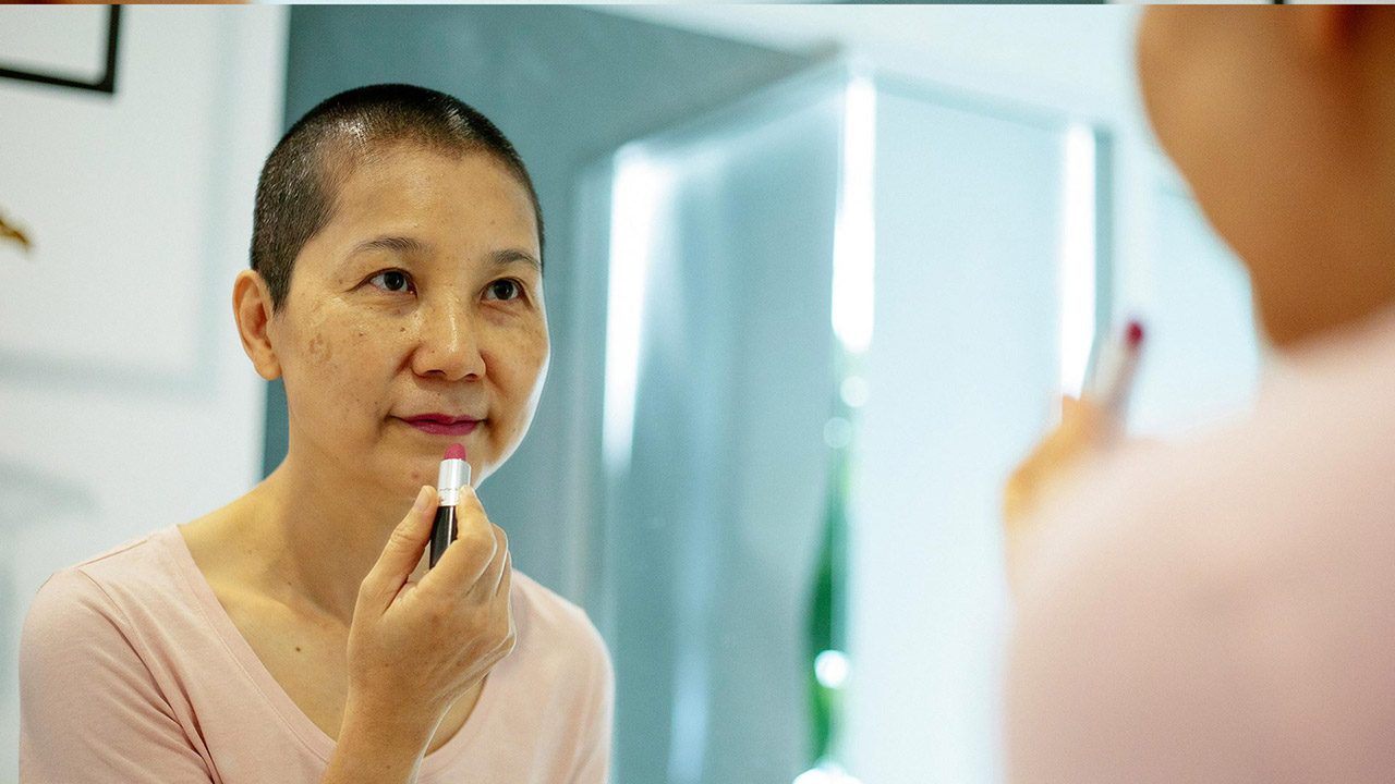 A middle-aged woman with a shaved head applying lipstick in the mirror