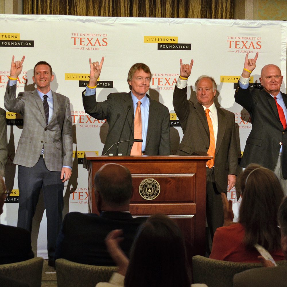 Four men at podium raising their hands in a "Hook 'em Horns" gesture