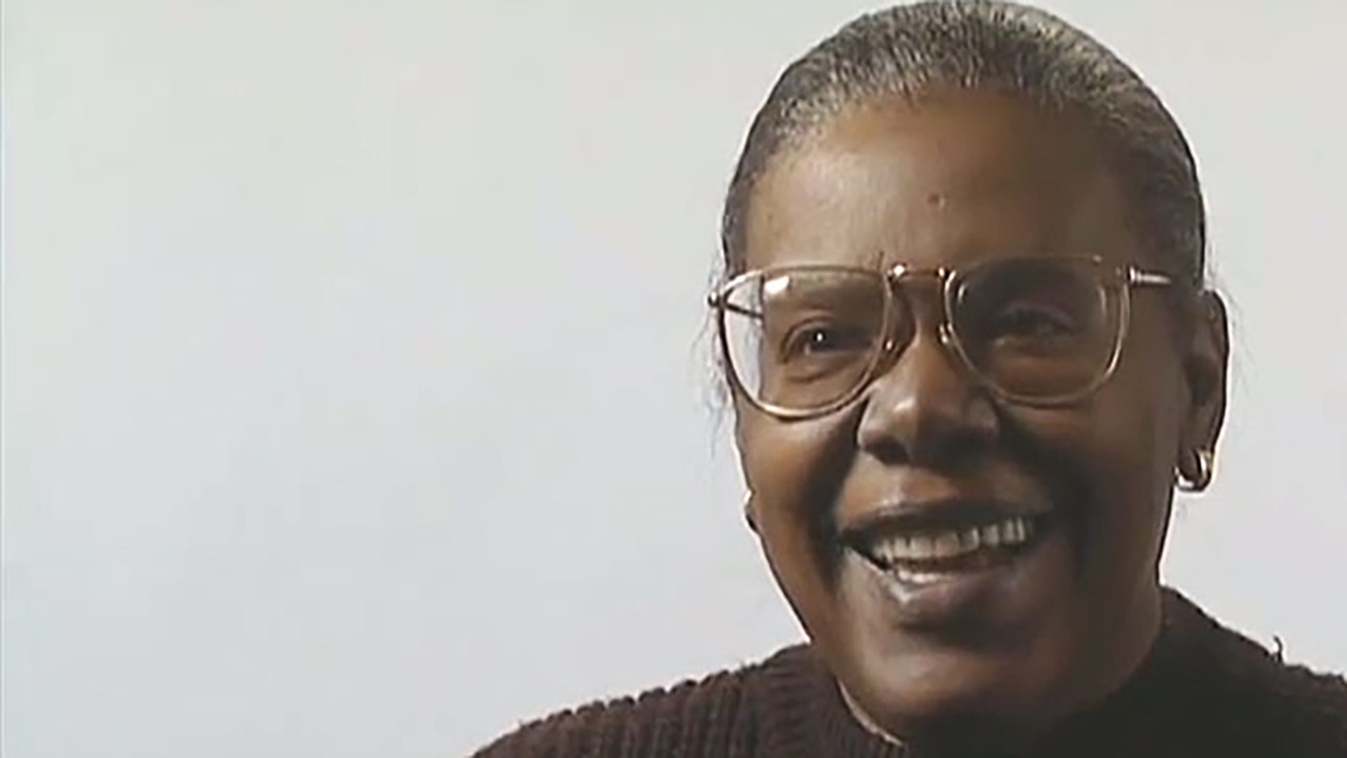 A smiling middle-aged woman wearing glasses and a brown sweater is interviewed against a white background.