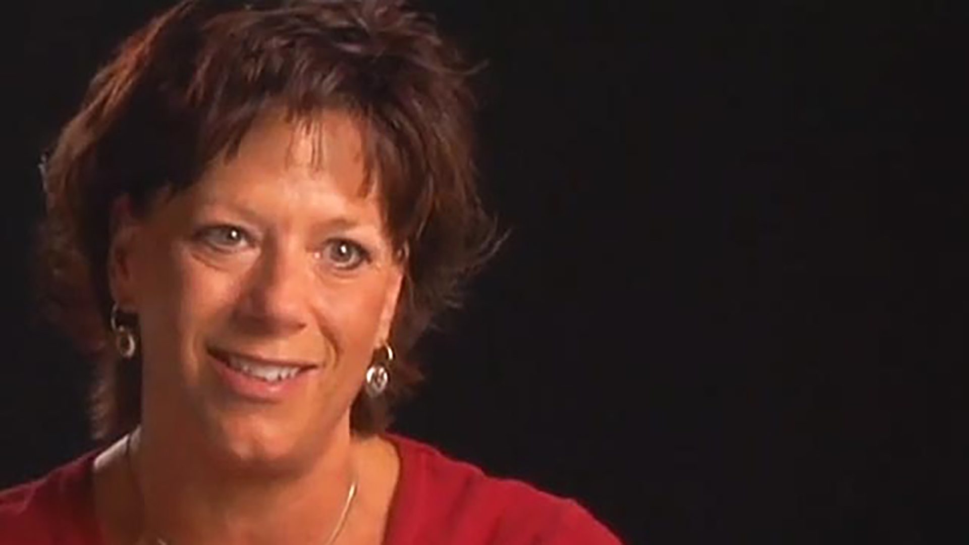 A middle-aged woman with short hear wears a red shirt and is interviewed against a black background.
