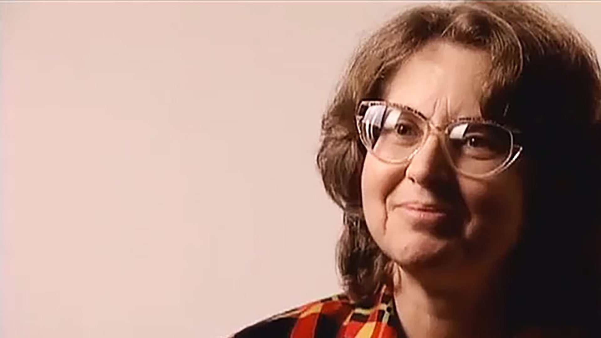 A middle-aged woman wearing glasses and a plaid shirt is interviewed against a neutral color background.