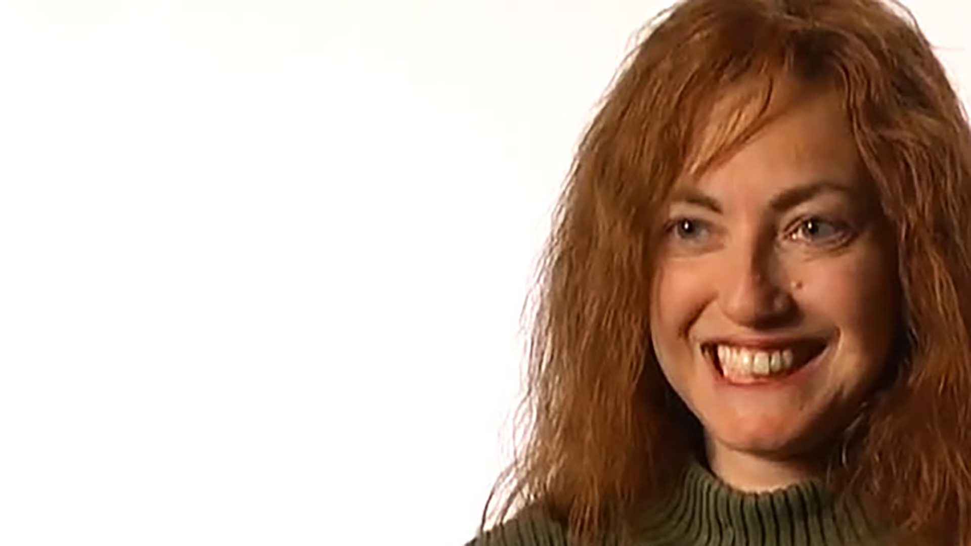 A young adult woman with red hair wears an olive green sweater and is interviewed against a white background.