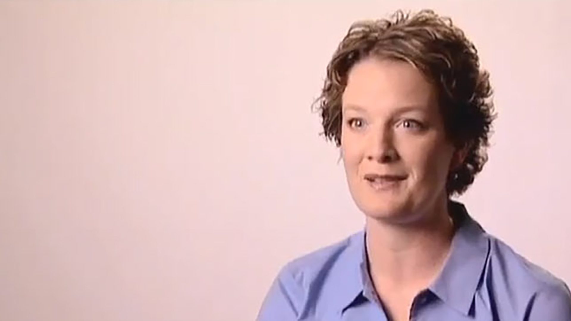 A woman with short curly hair wearing a light blue button-down shirt is interviewed against a light background.