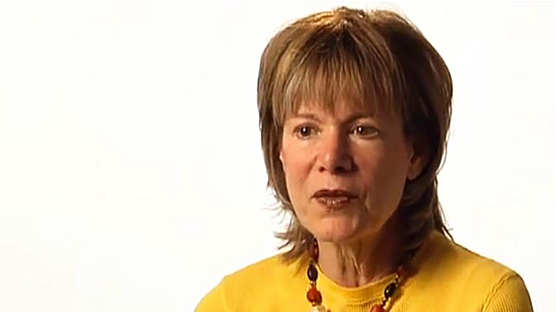 A middle-aged woman wearing a yellow sweater is interviewed against a white background