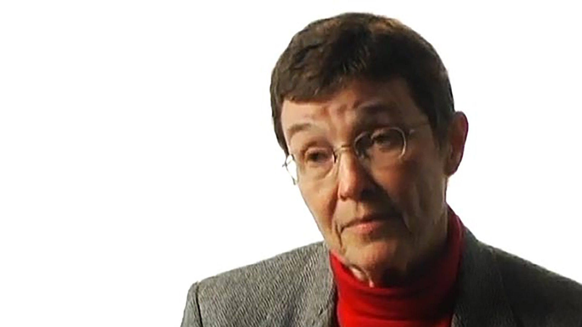 An older woman with short hair wearing glasses, a red turtleneck, and a gray jacket is interviewed against a white background