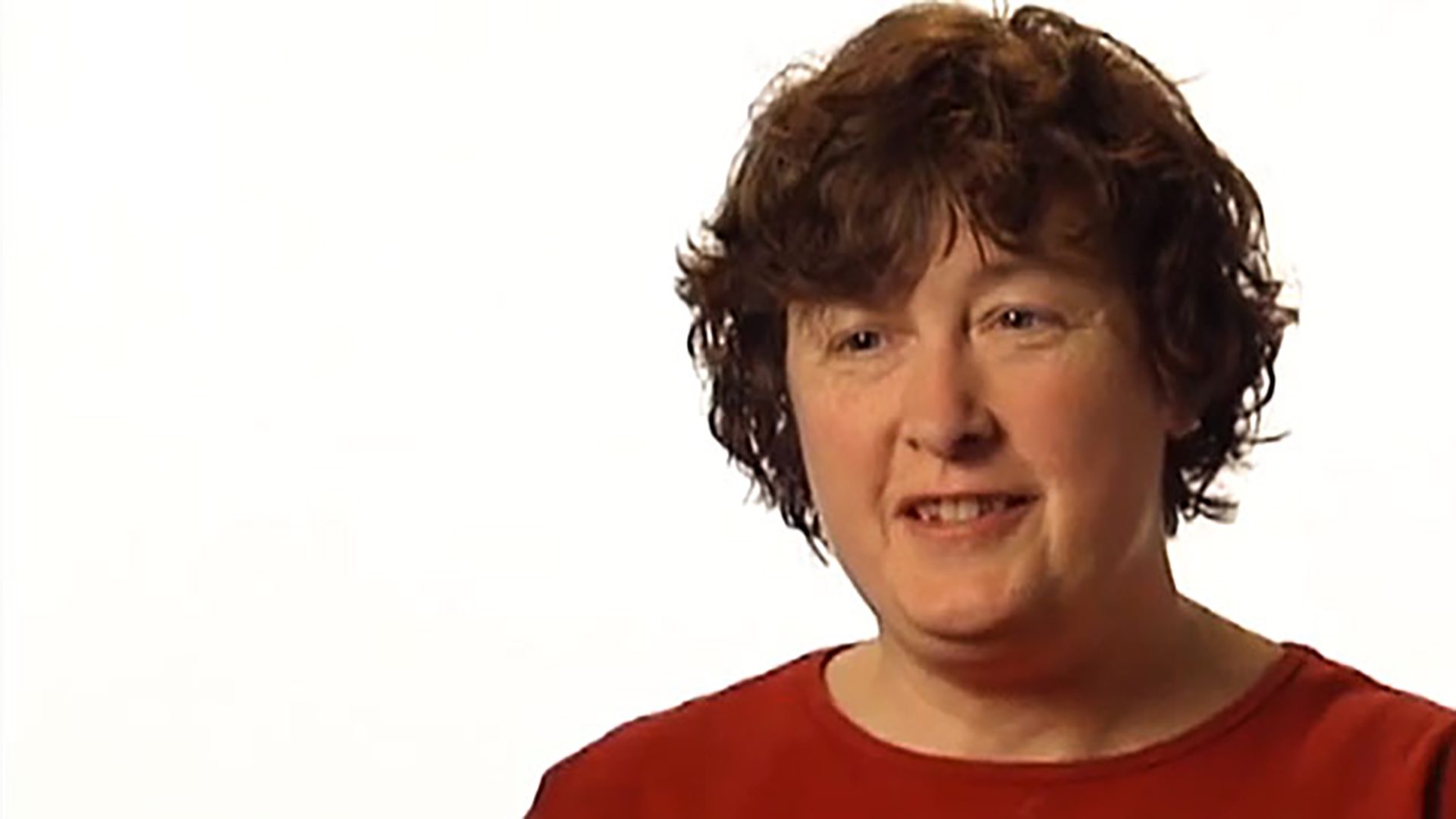 A middle-aged woman with short hair wearing a red shirt is interviewed against a white background.