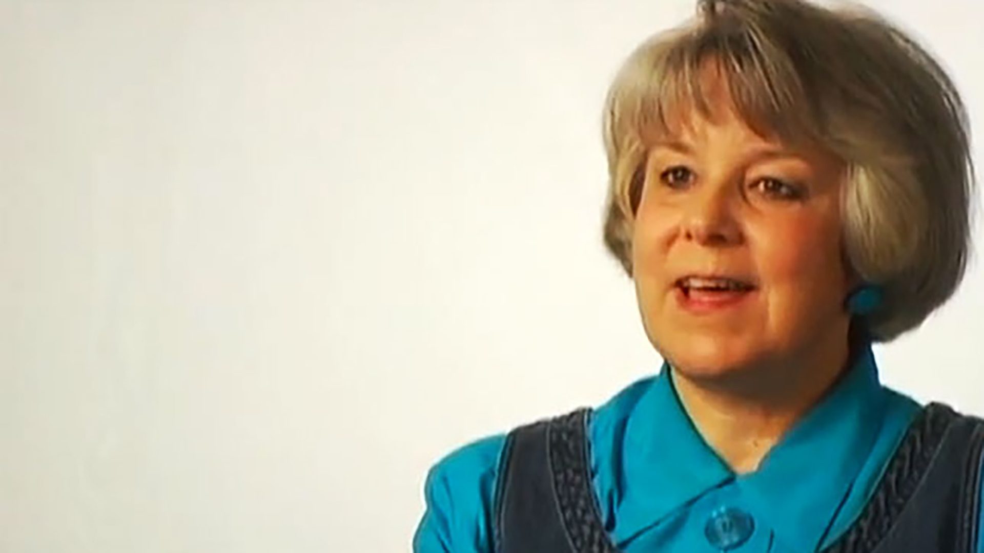A senior woman wearing a teal shirt is interviewed against a white background.