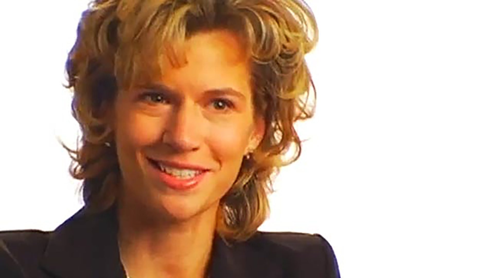 A woman with curly blonde hair wearing a blazer is interviewed against a white background.