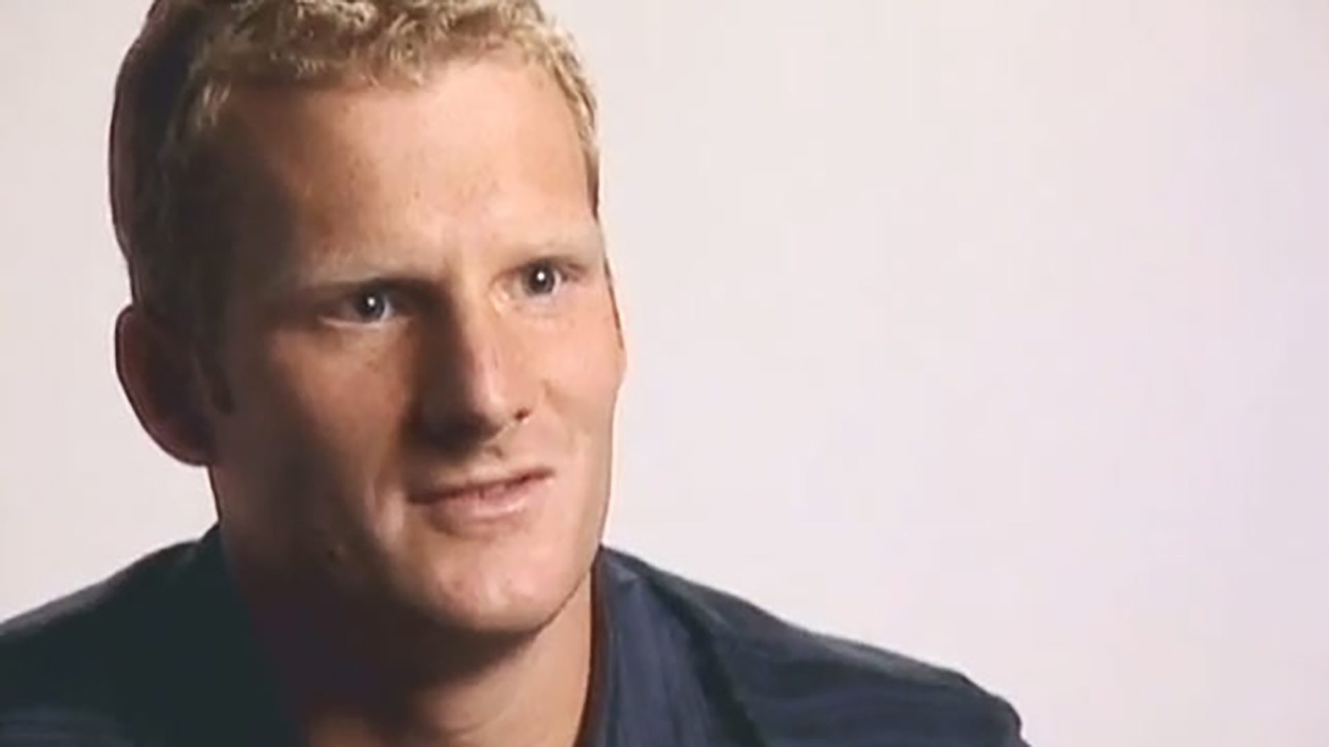 A blond man wearing a dark shirt is interviewed against a white background