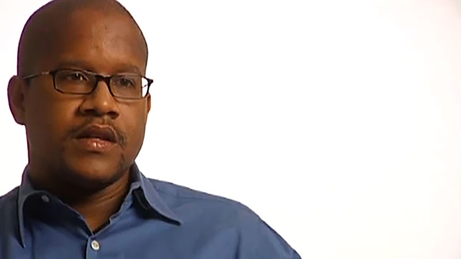 A bald adult man wearing glasses and a blue button-up shirt is interviewed against a white background.