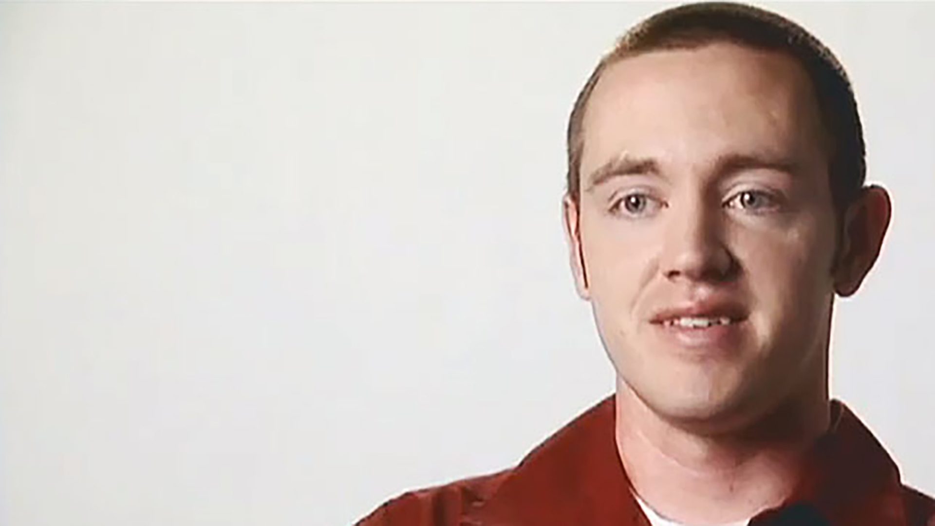 A young man with short hair wearing a red collared shirt is interviewed against a white background