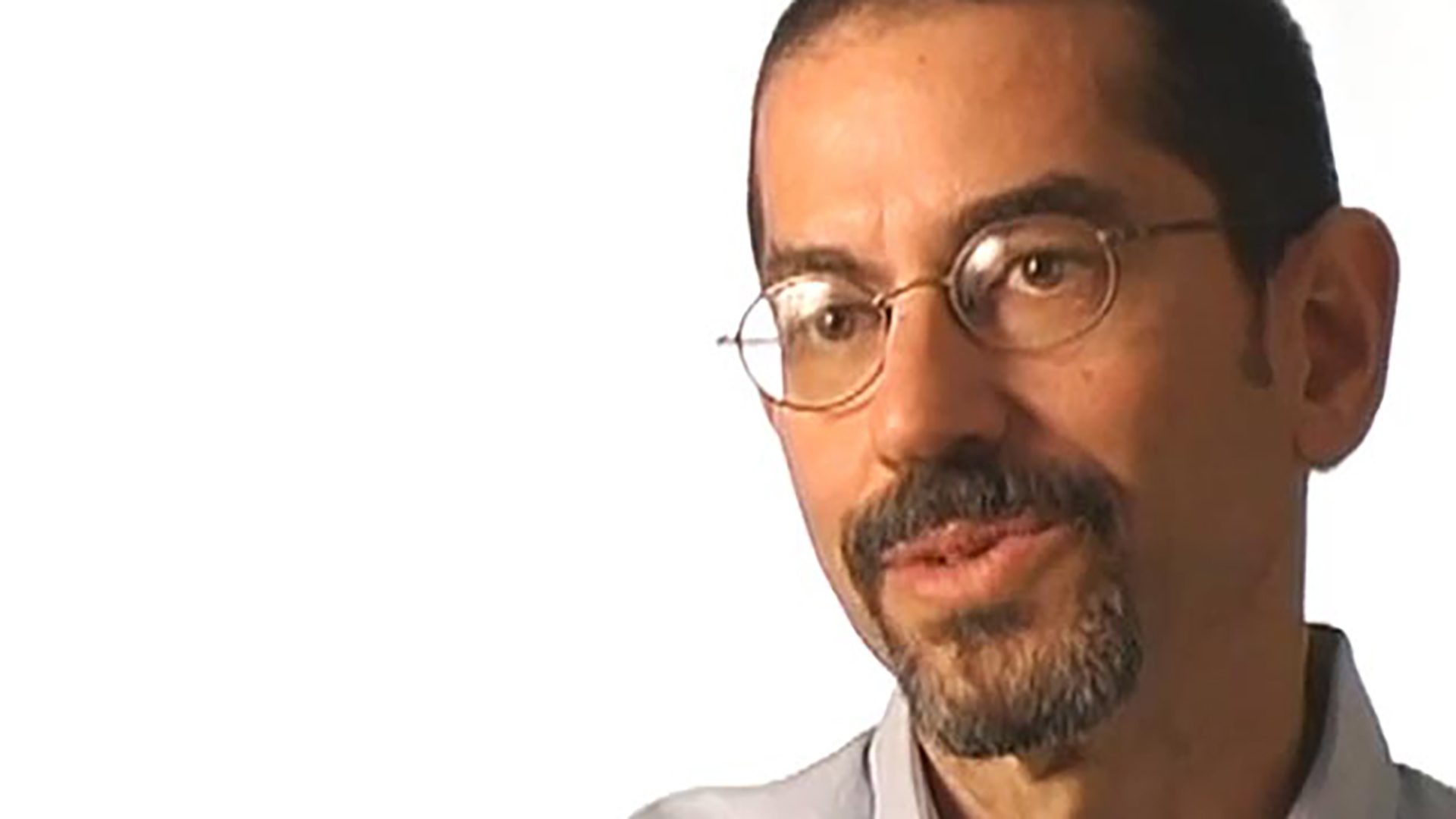 An adult man with a buzz cut hairstyle, facial hair, and glasses is interviewed against a white background.