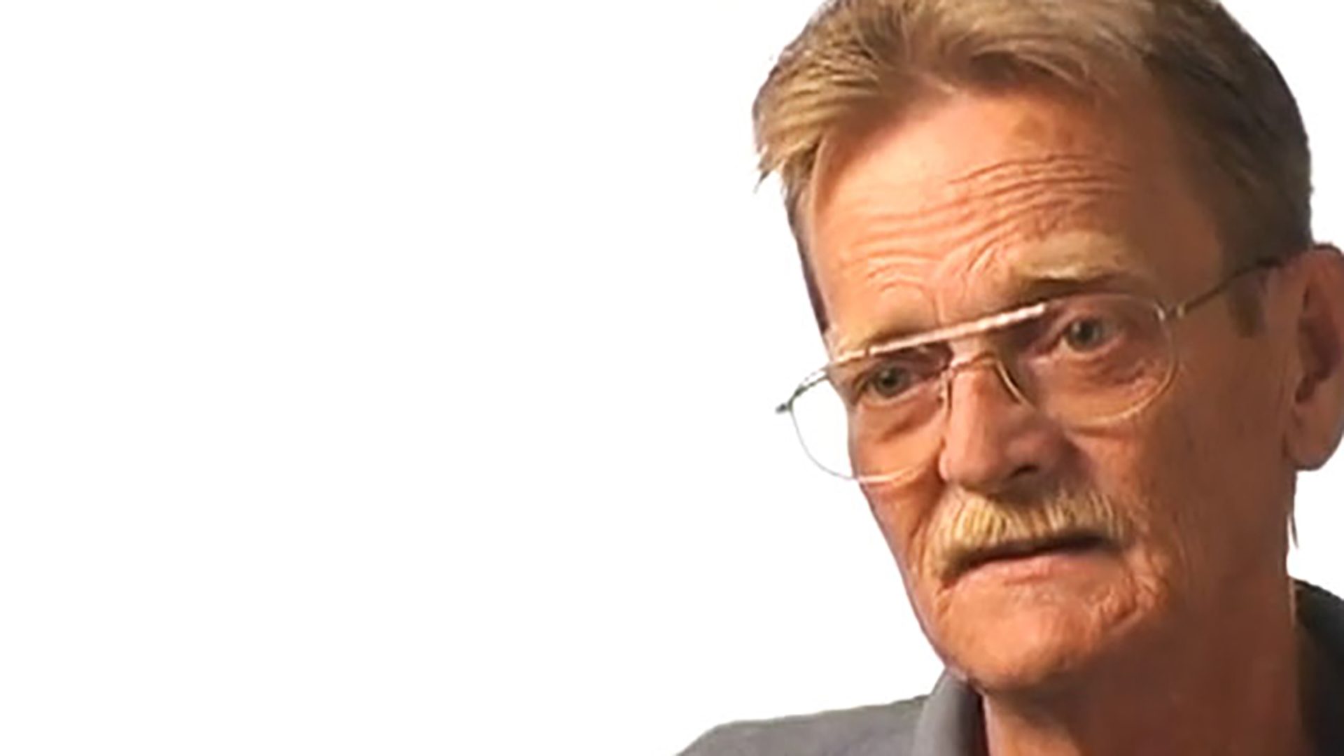 A senior man with glasses and a mustache is interviewed against a white background.