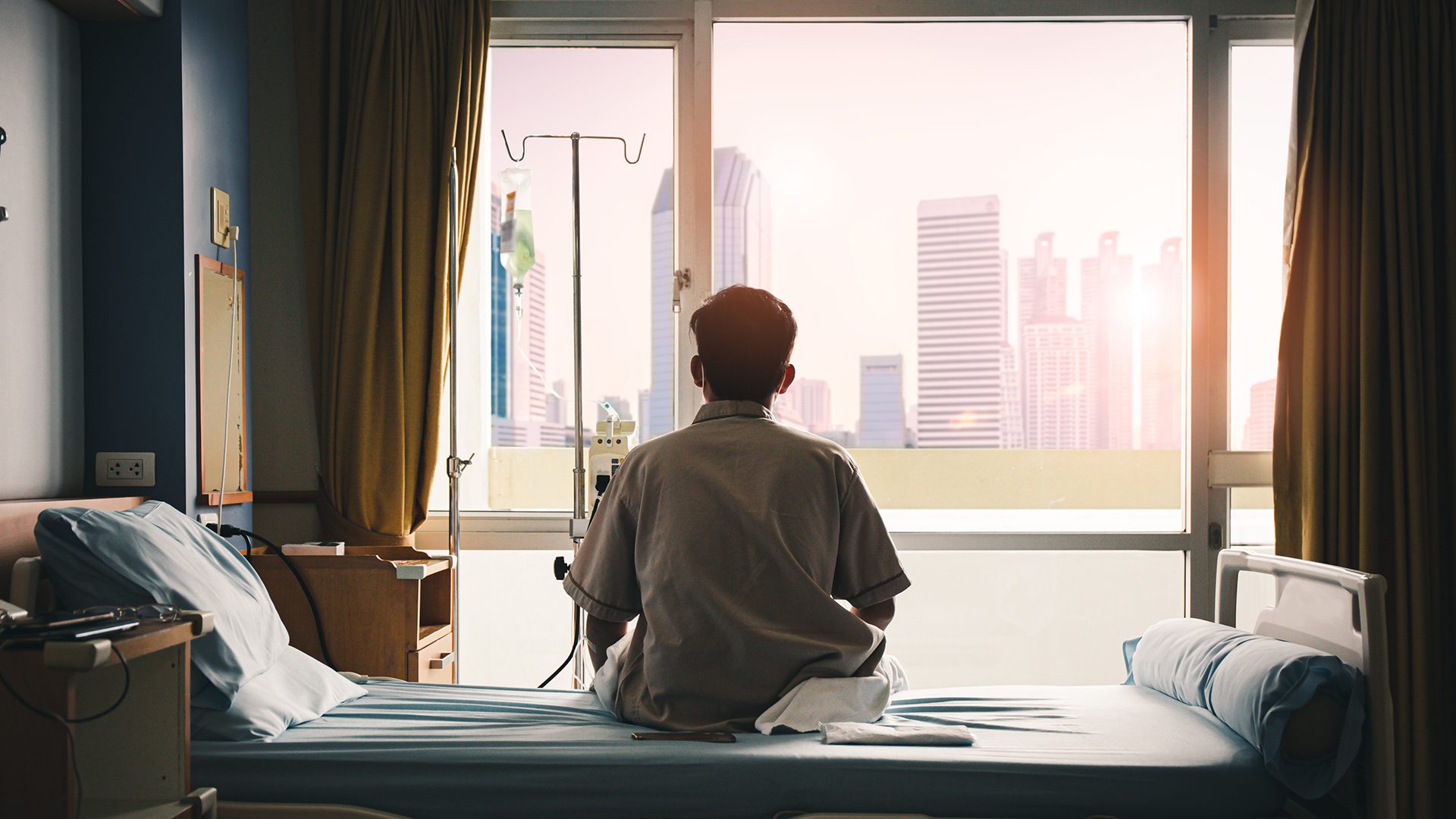 A man with his back to the camera looks out the window of his hospital room