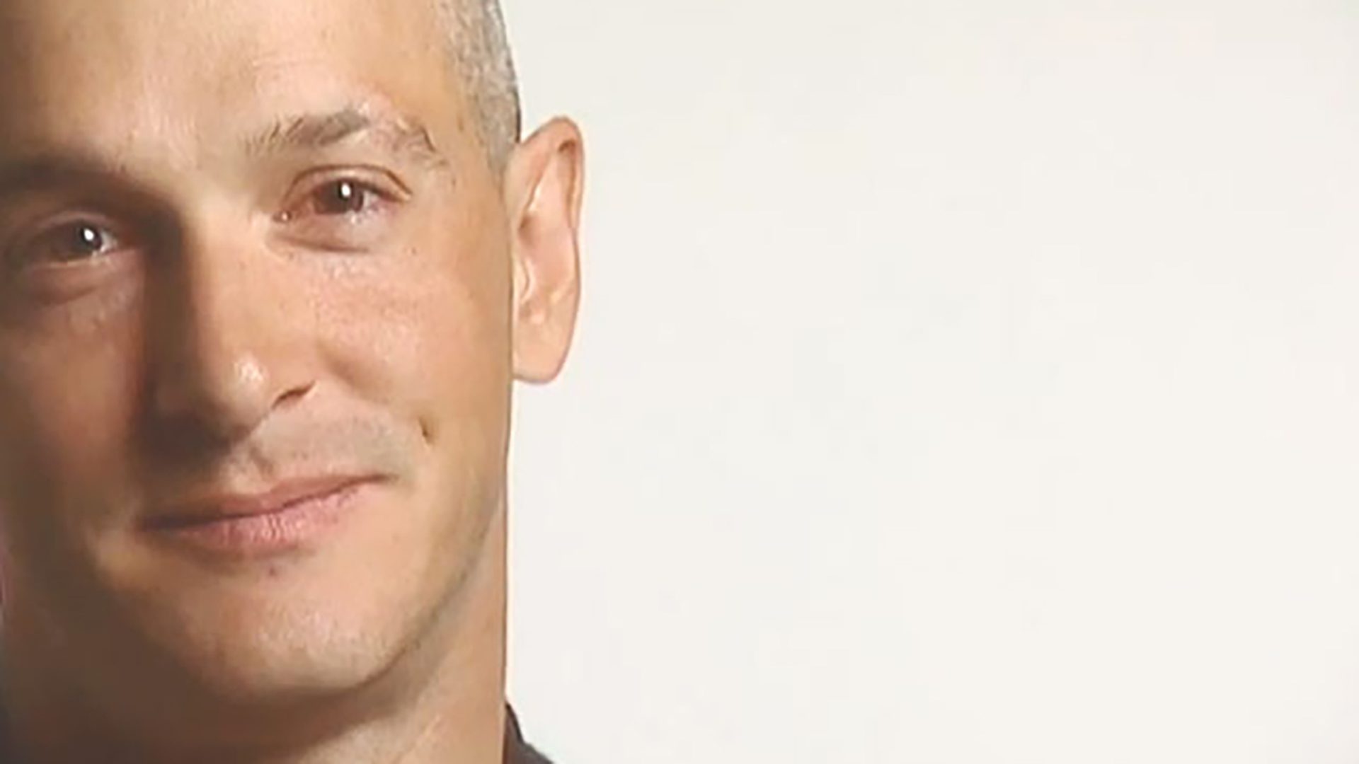 A close-up portrait of a man with a buzz cut hairstyle against a white background