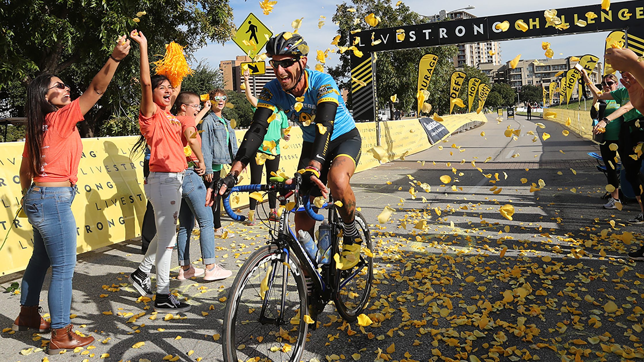 Man on bike crossing finish line of race while onlookers cheer and throw yellow rose petals