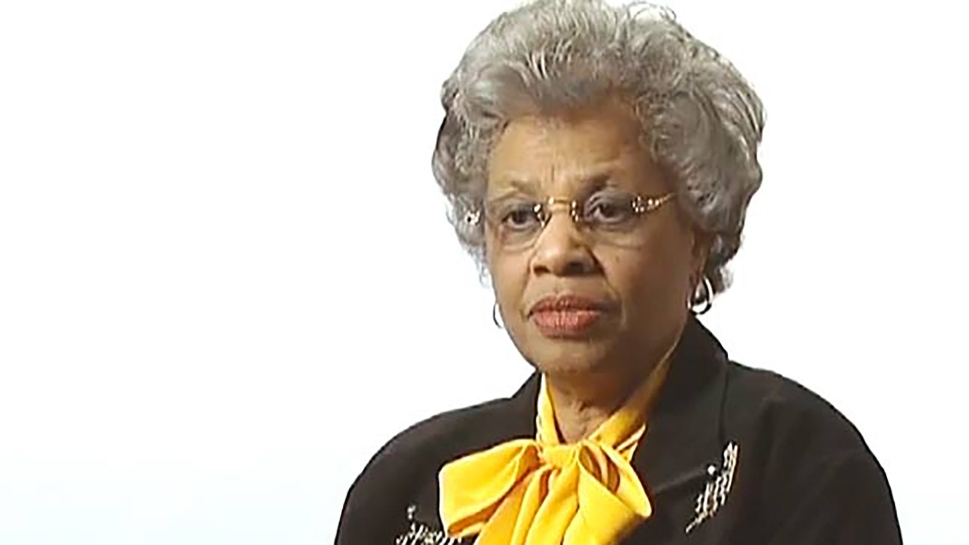 A senior woman with curly gray hair wearing glasses and a yellow necktie is interviewed against a white background.