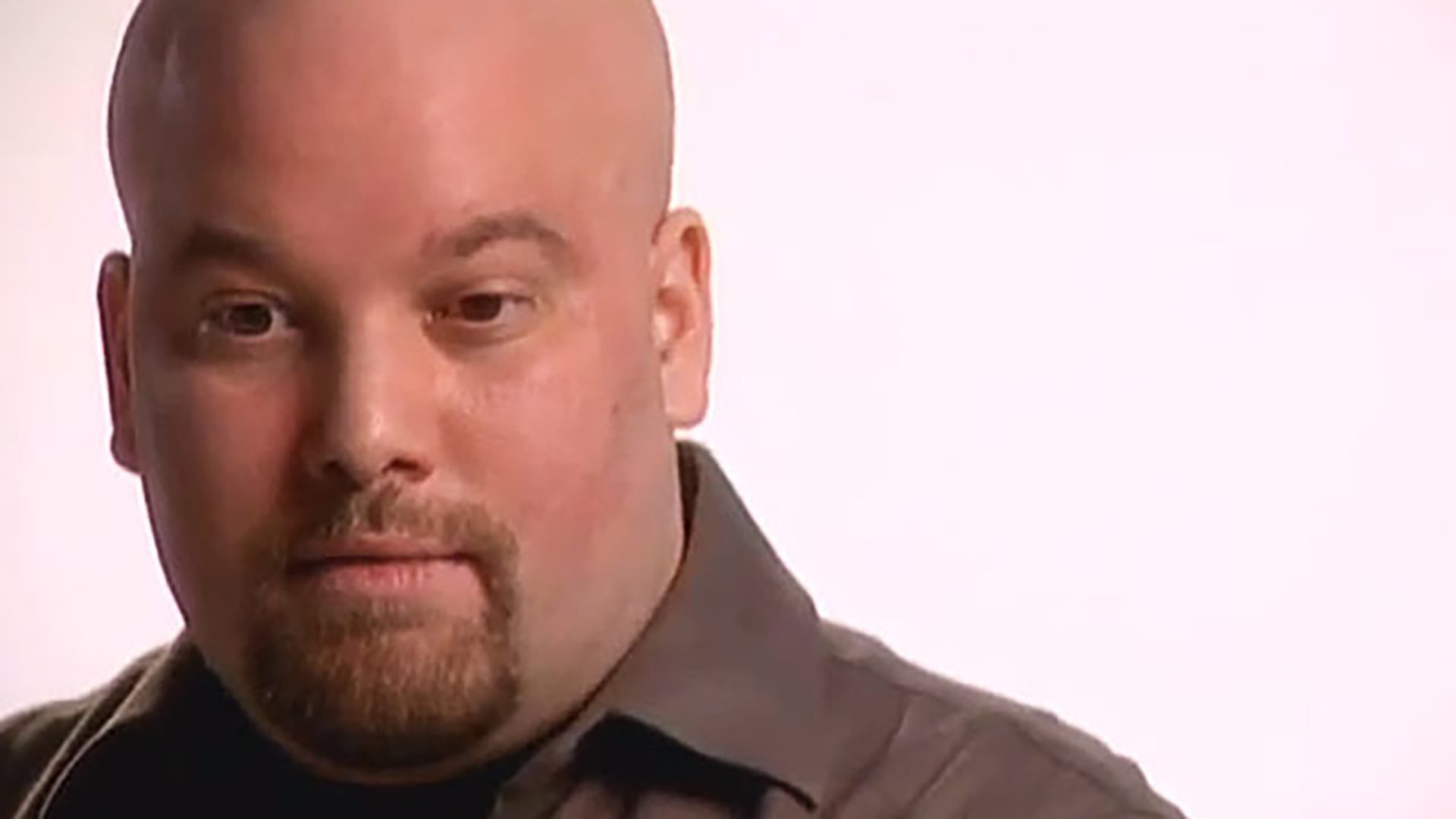 A bald man with a goatee wearing a brown collared shirt is interviewed against a white background