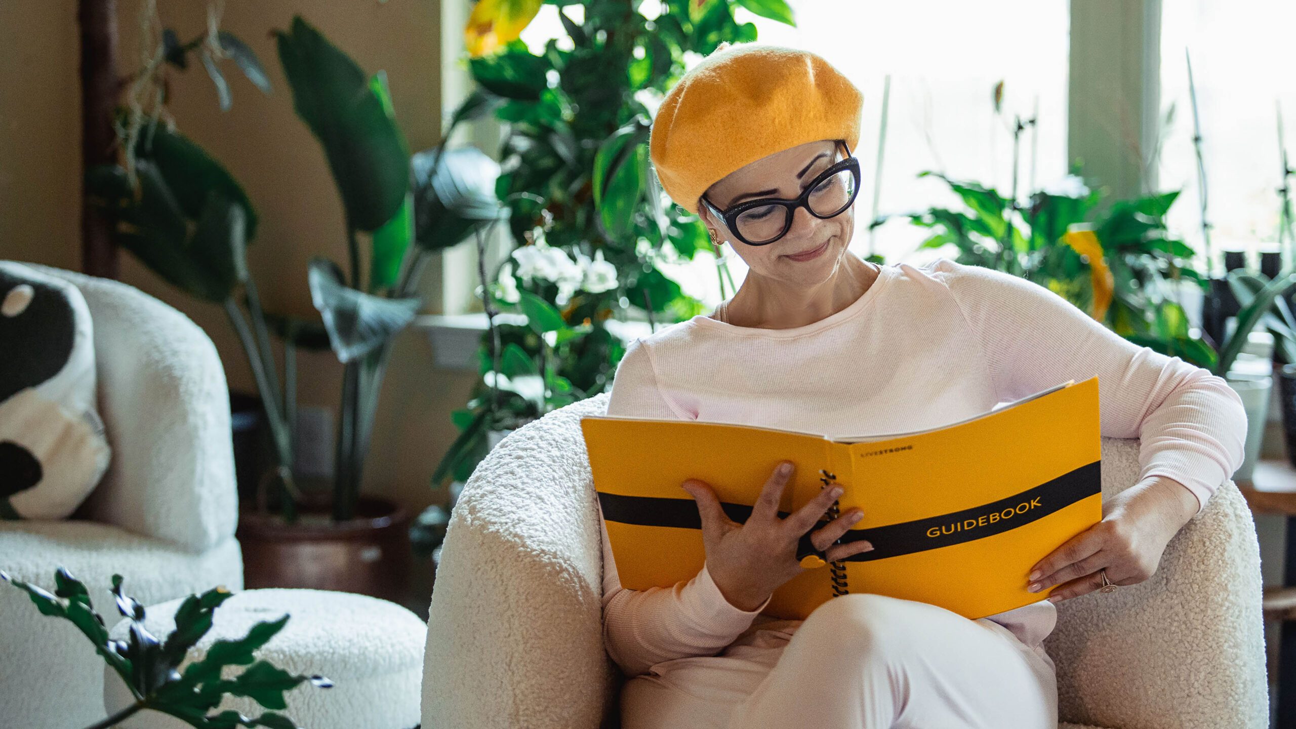 A woman in glasses and a yellow hat sitting in a chair and reading a yellow book