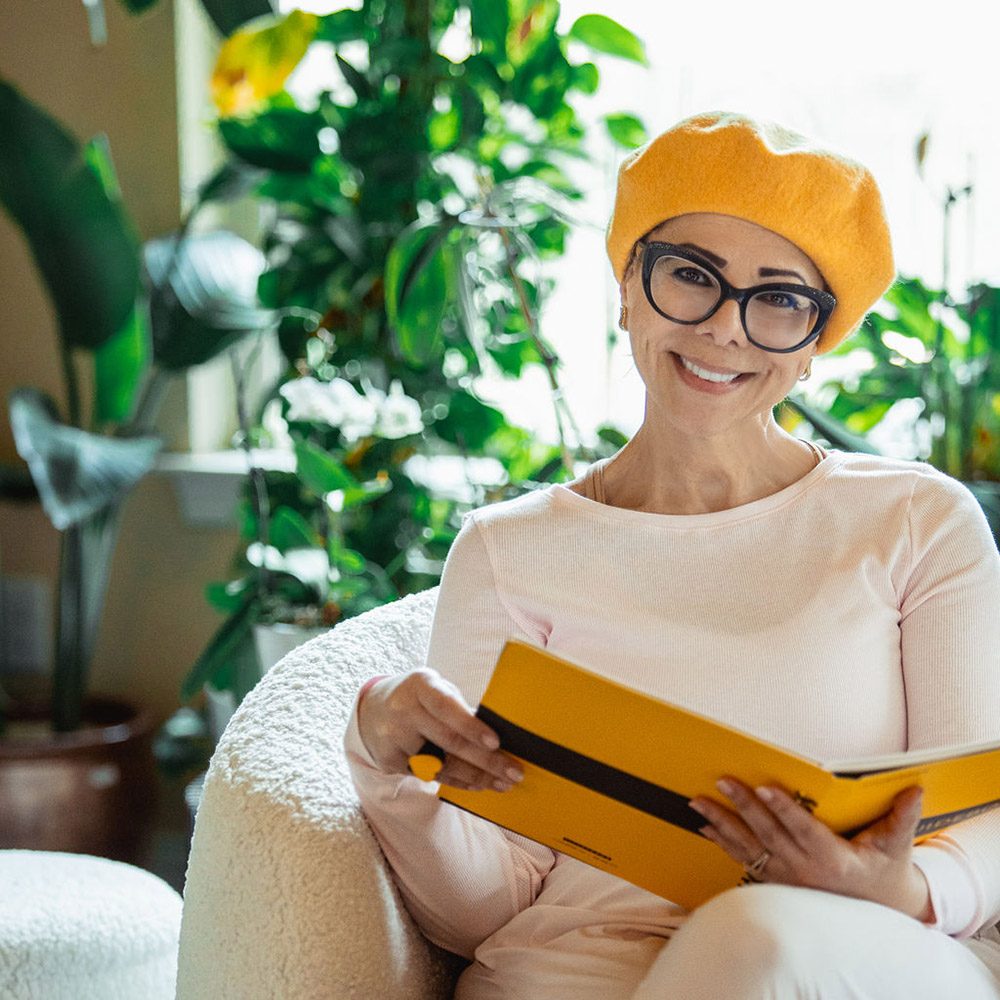 A woman wearing glasses and a yellow hat sits in a chair and reads a yellow book.