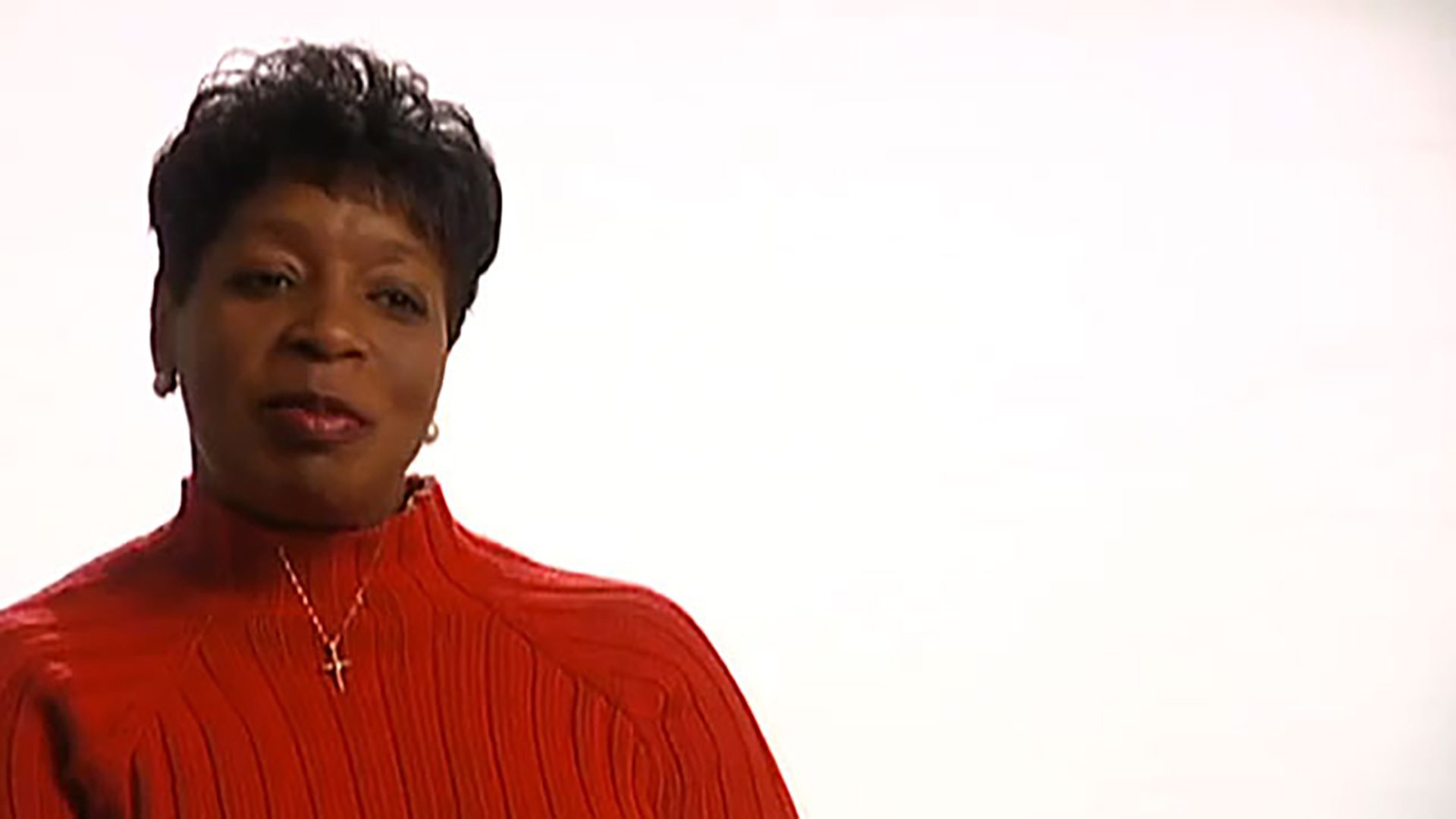A middle aged woman with short black hair wearing a red turtleneck is interviewed against a white background