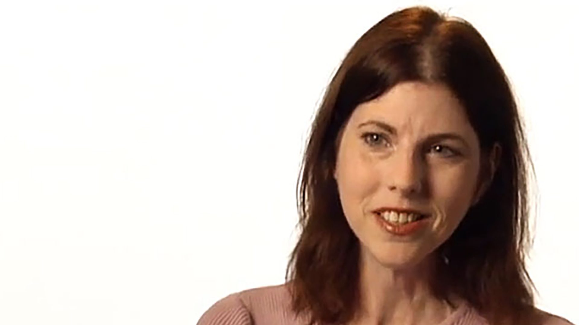 An adult woman with shoulder-length brown hair wears a pink shirt and is interviewed against a white background.