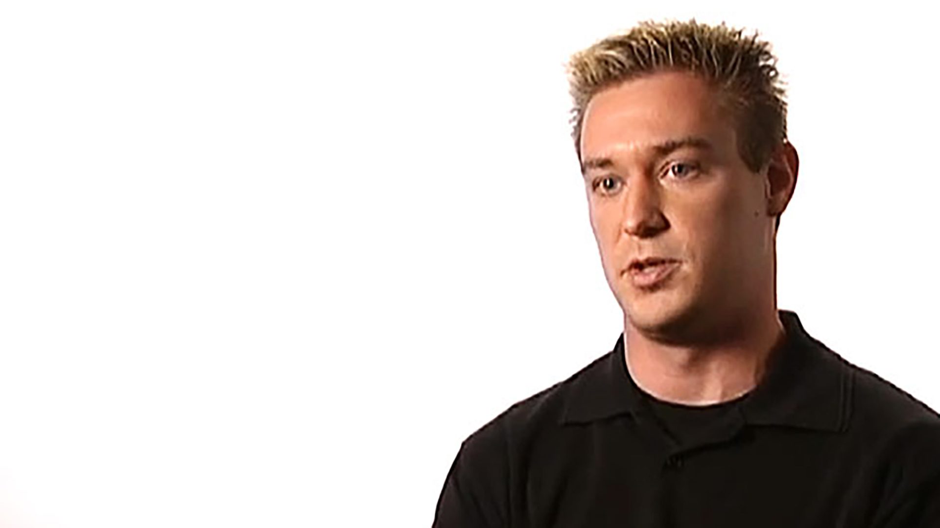 A man with spiked blond hair and a black shirt is interviewed against a white background
