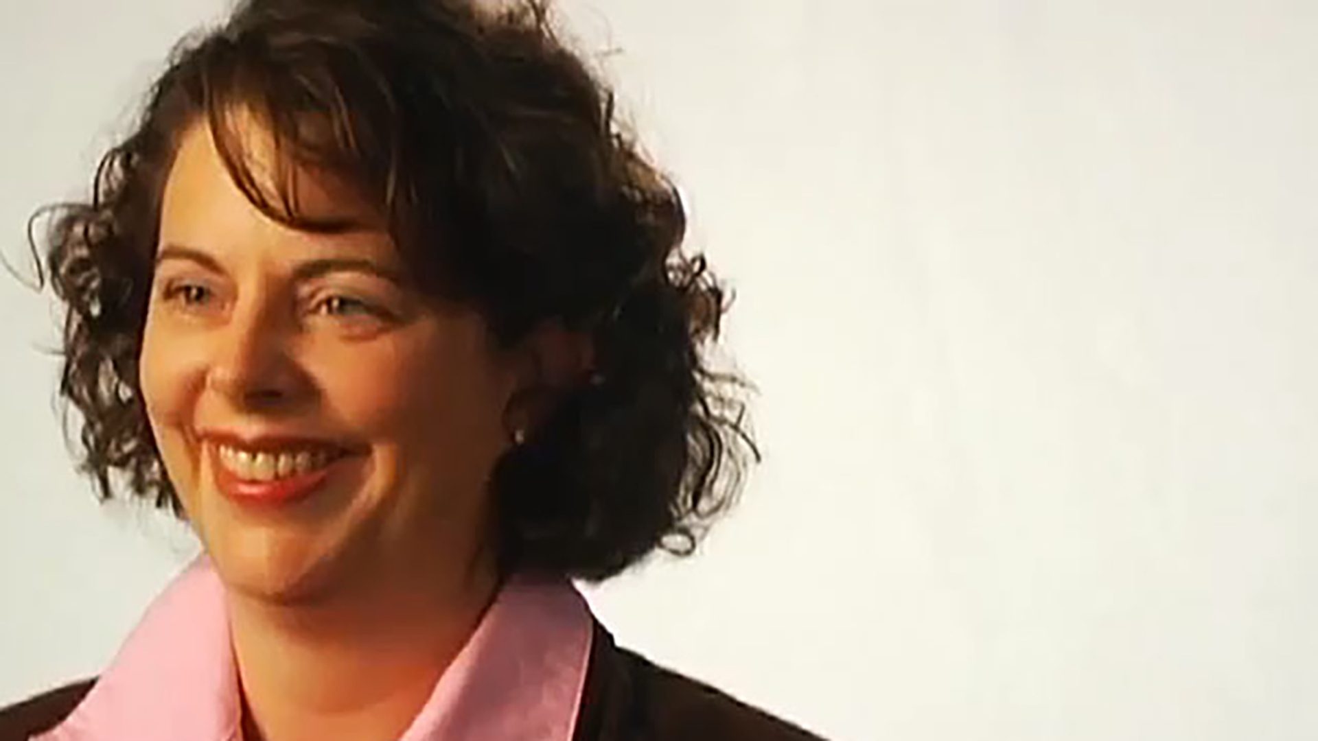 A woman with short, dark, curly hair wears a light pink and brown shirt and is interviewed against a white background.
