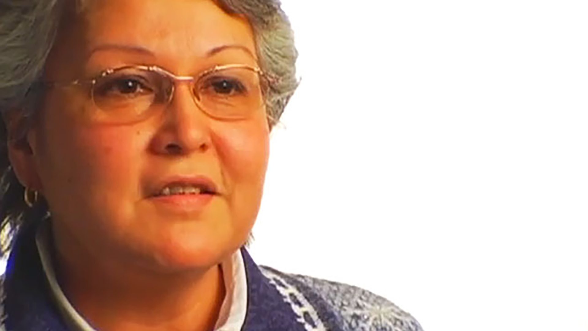 A senior woman with short gray hair and glasses is interviewed against a white background