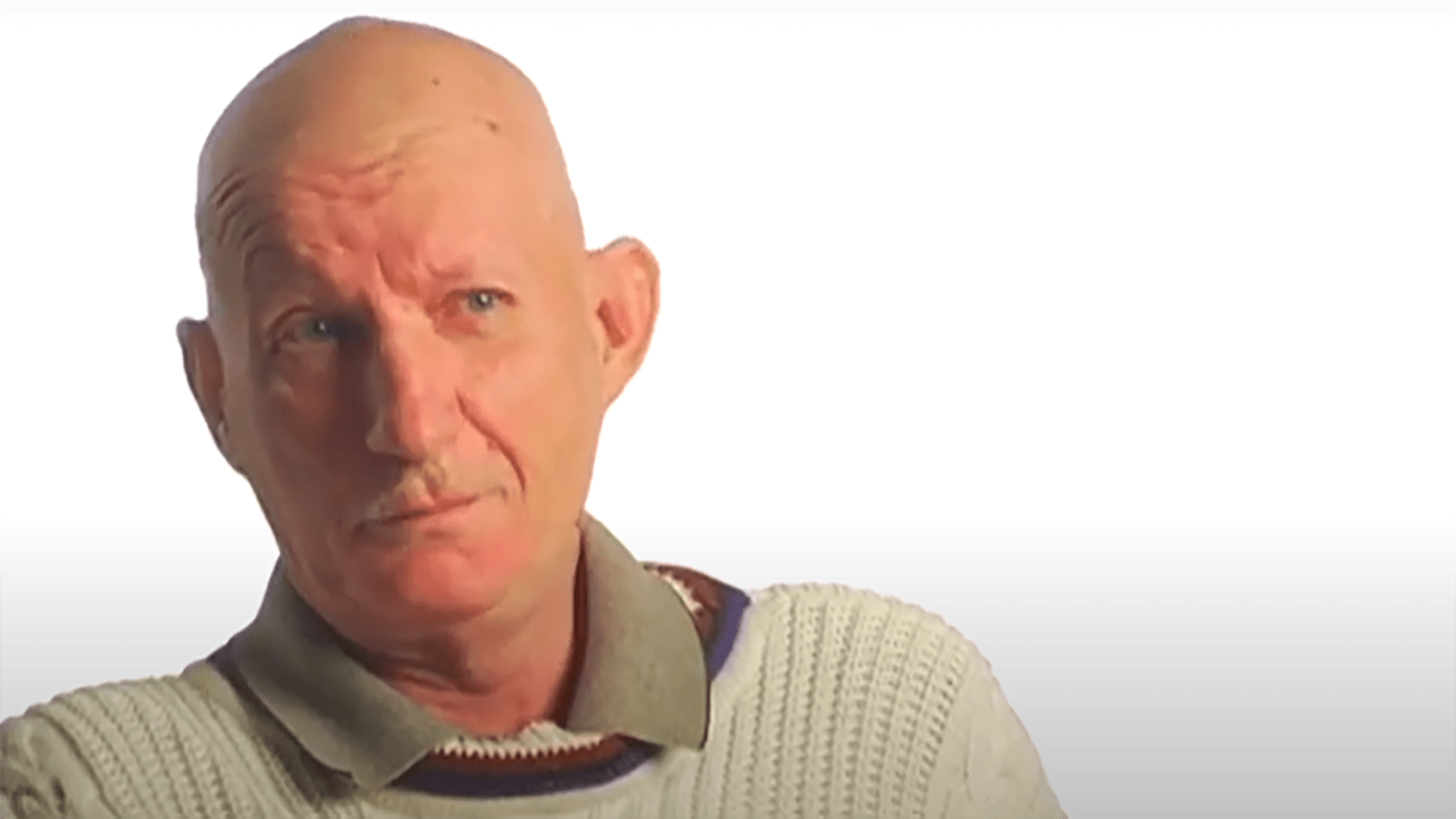 A bald middle aged gentleman is interviewed wearing a white sweater