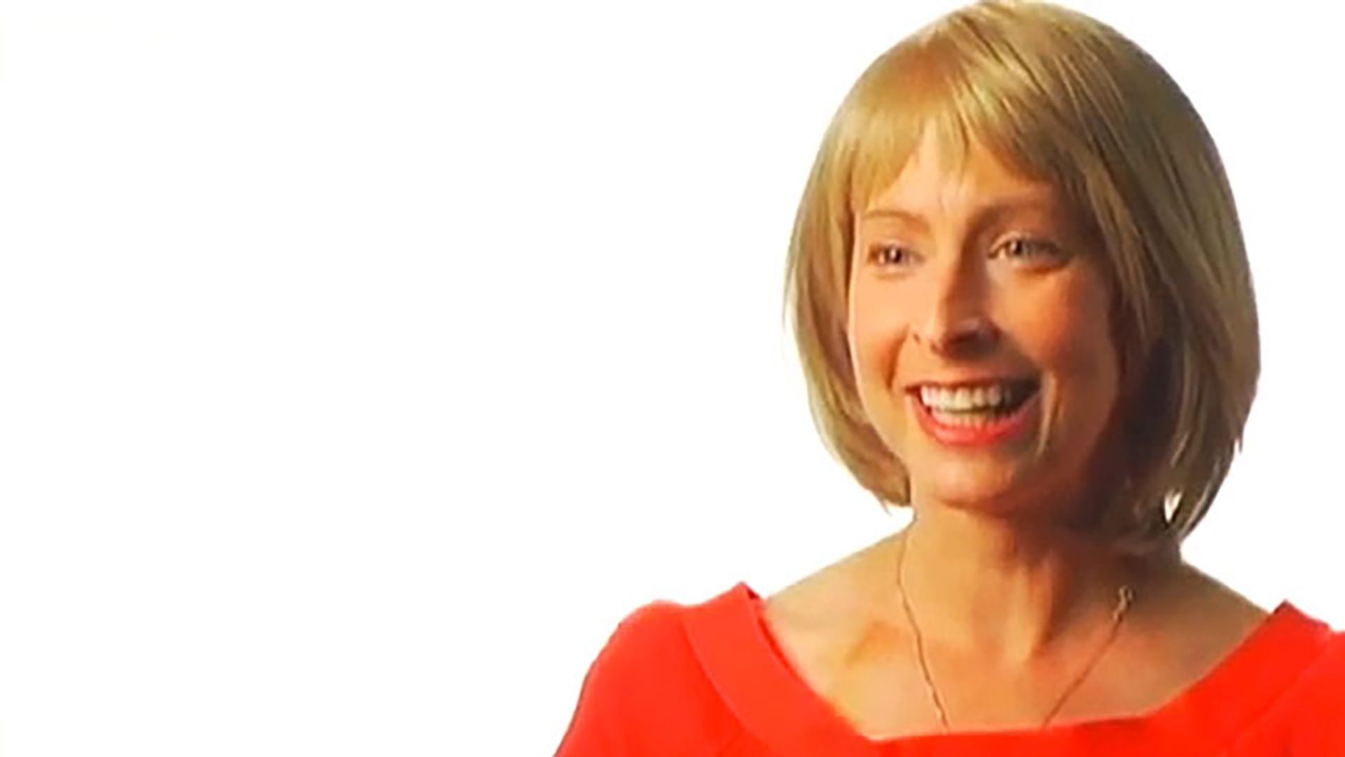 A smiling blonde woman wearing a red shirt is interviewed against a white background