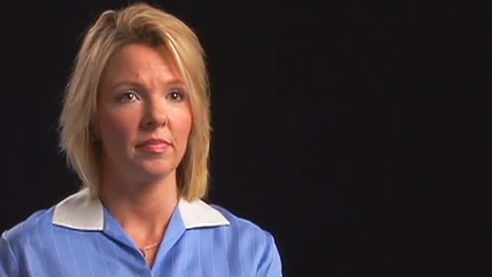 A woman with blonde shoulder-length hair and a light blue collared shirt is interviewed against a black background.