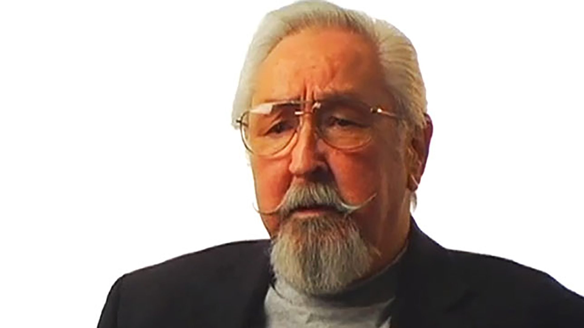 A senior man with glasses and facial hair is interviewed against a white background.