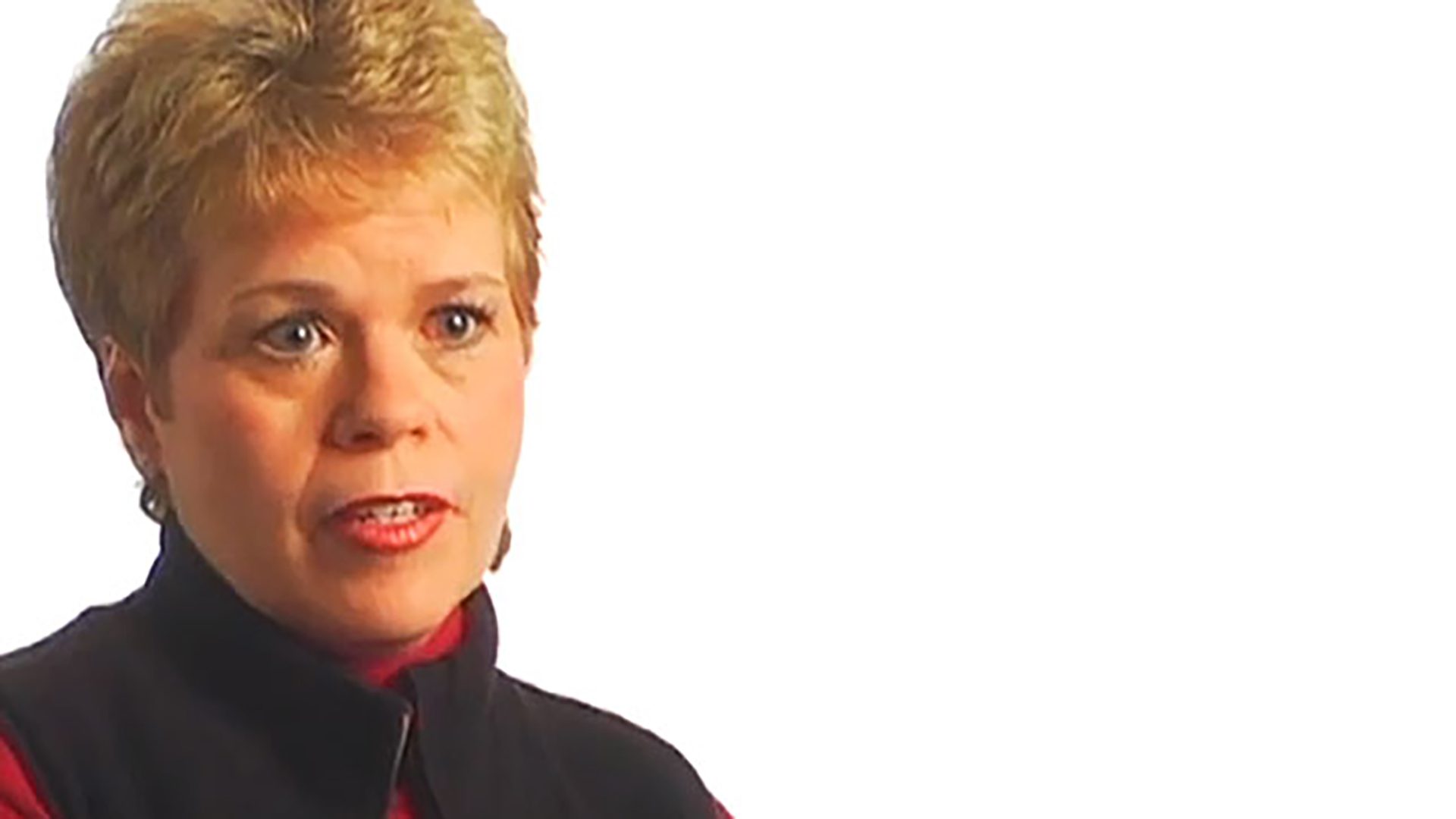 A middle-aged woman with short blonde hair wears a red turtleneck and black vest. She is interviewed against a white background.