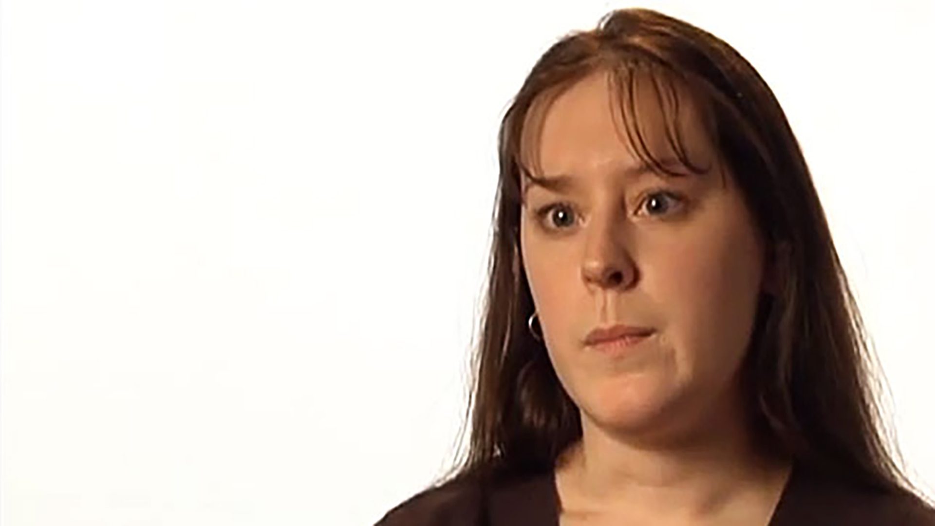 A young woman with dark hair and a dark shirt is interviewed against a white background.