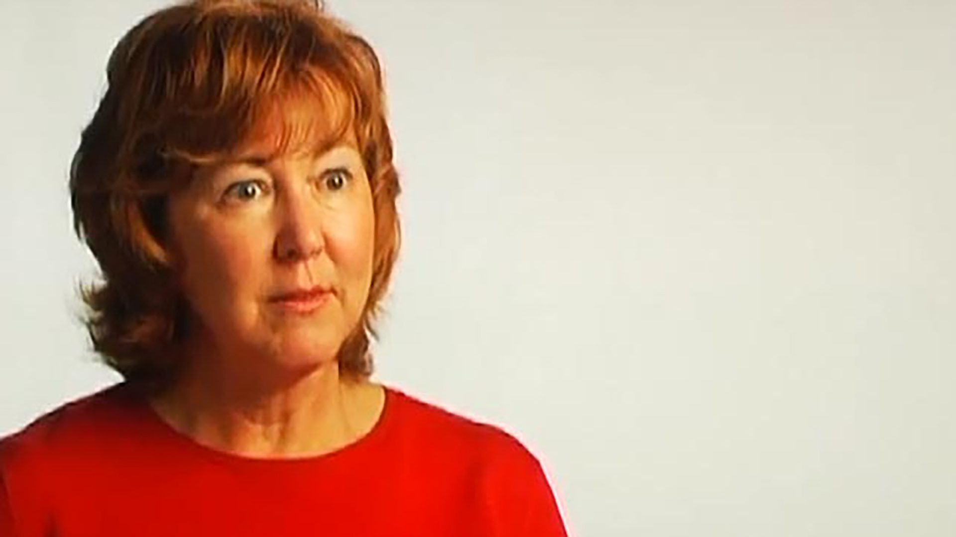 A middle-aged woman with red hair wears a red shirt and is interviewed against a white background.
