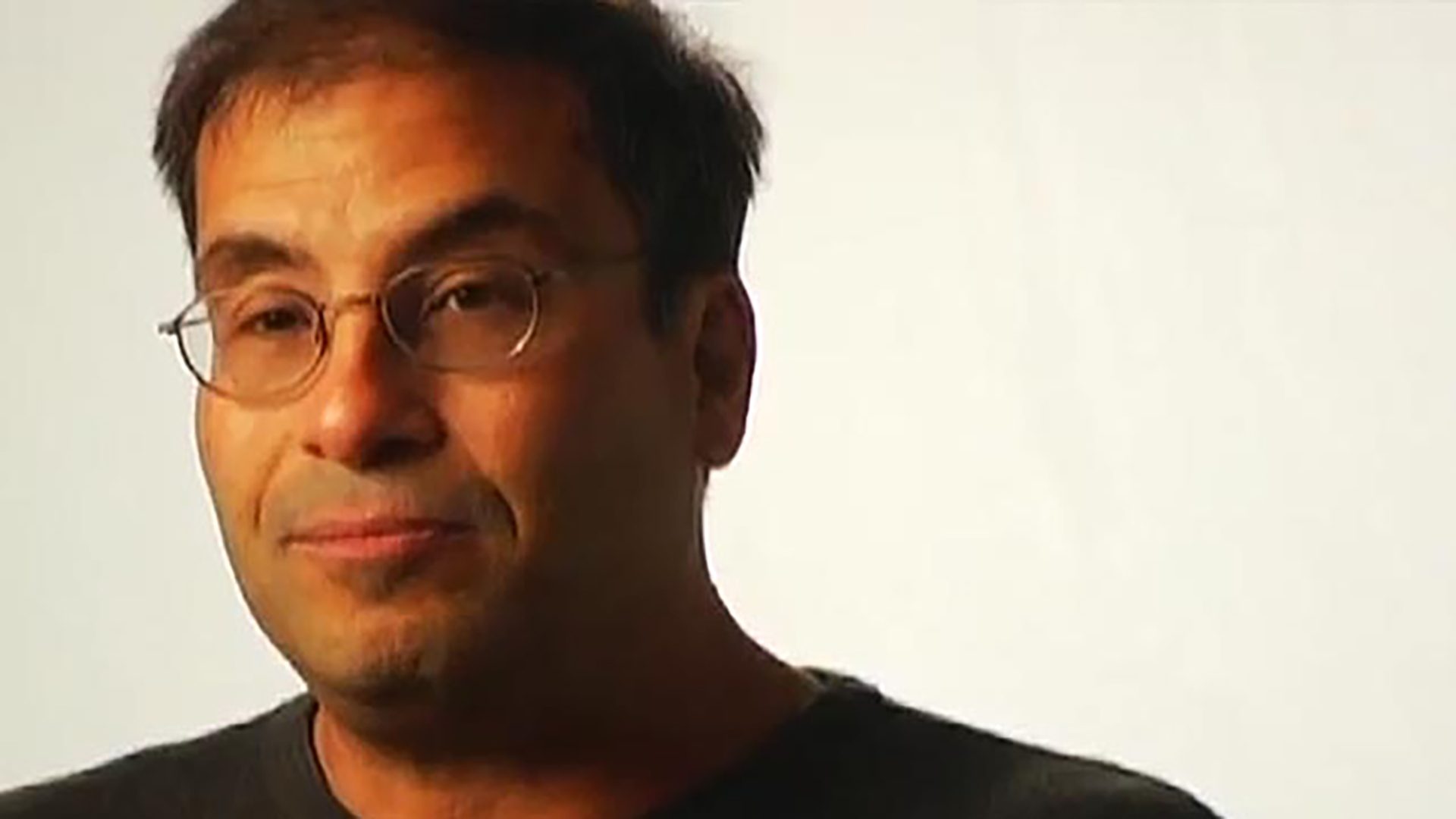 A middle-aged man wearing glasses and a black t-shirt is interviewed against a white background