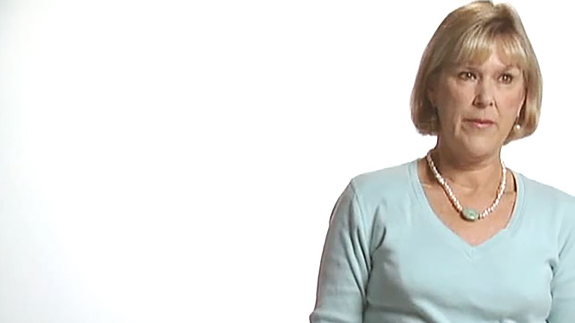 A middle-aged woman with a blonde bob haircut wears a light blue long-sleeved shirt and is interviewed against a white background.