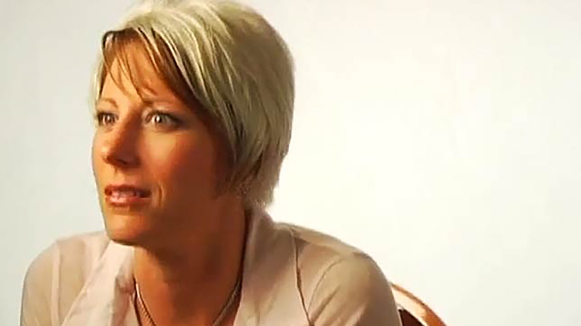A woman with short blonde hair wearing a white shirt is interviewed against a white background.