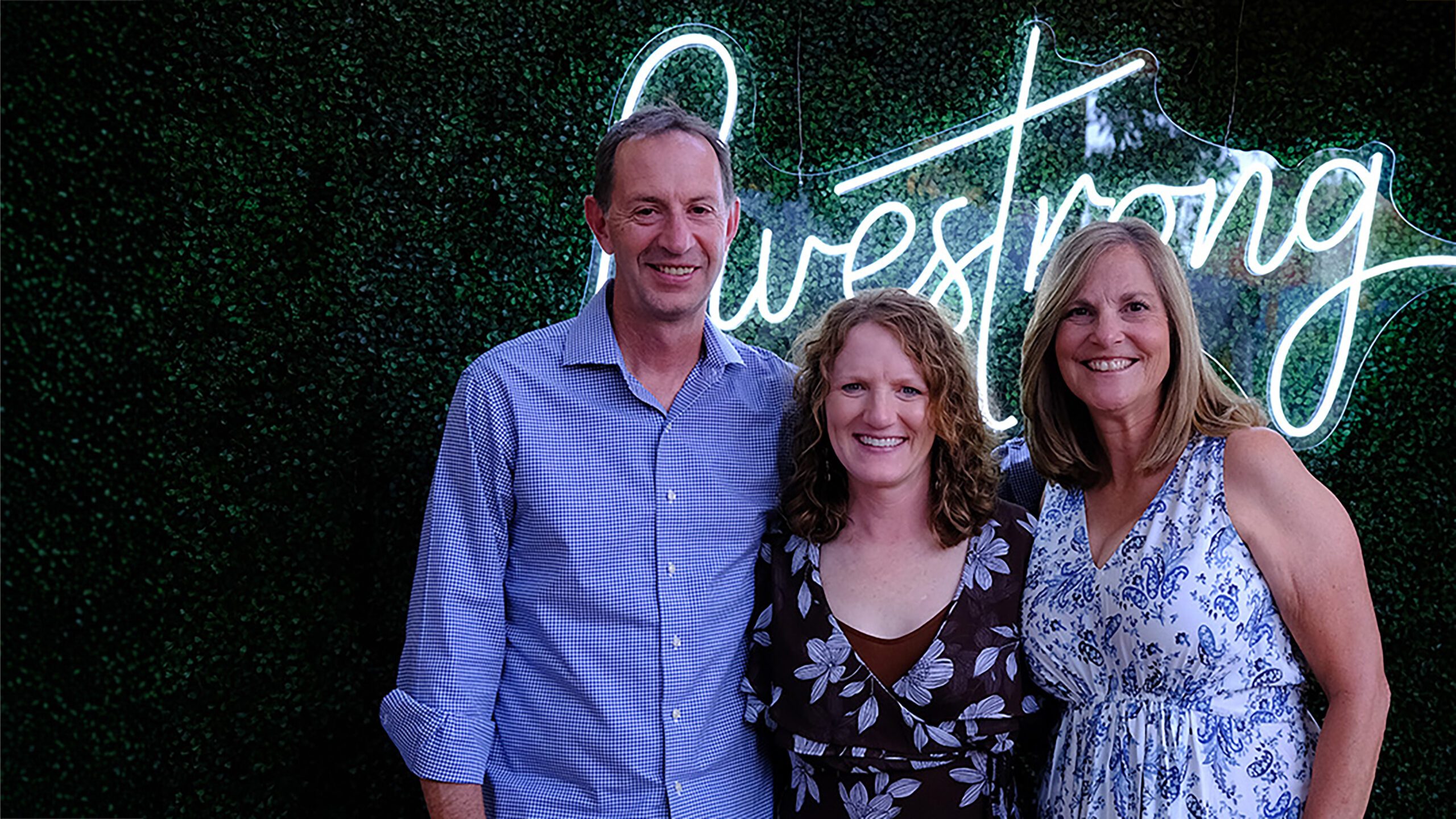 A man and two women standing in front of Livestrong neon sign and greenery