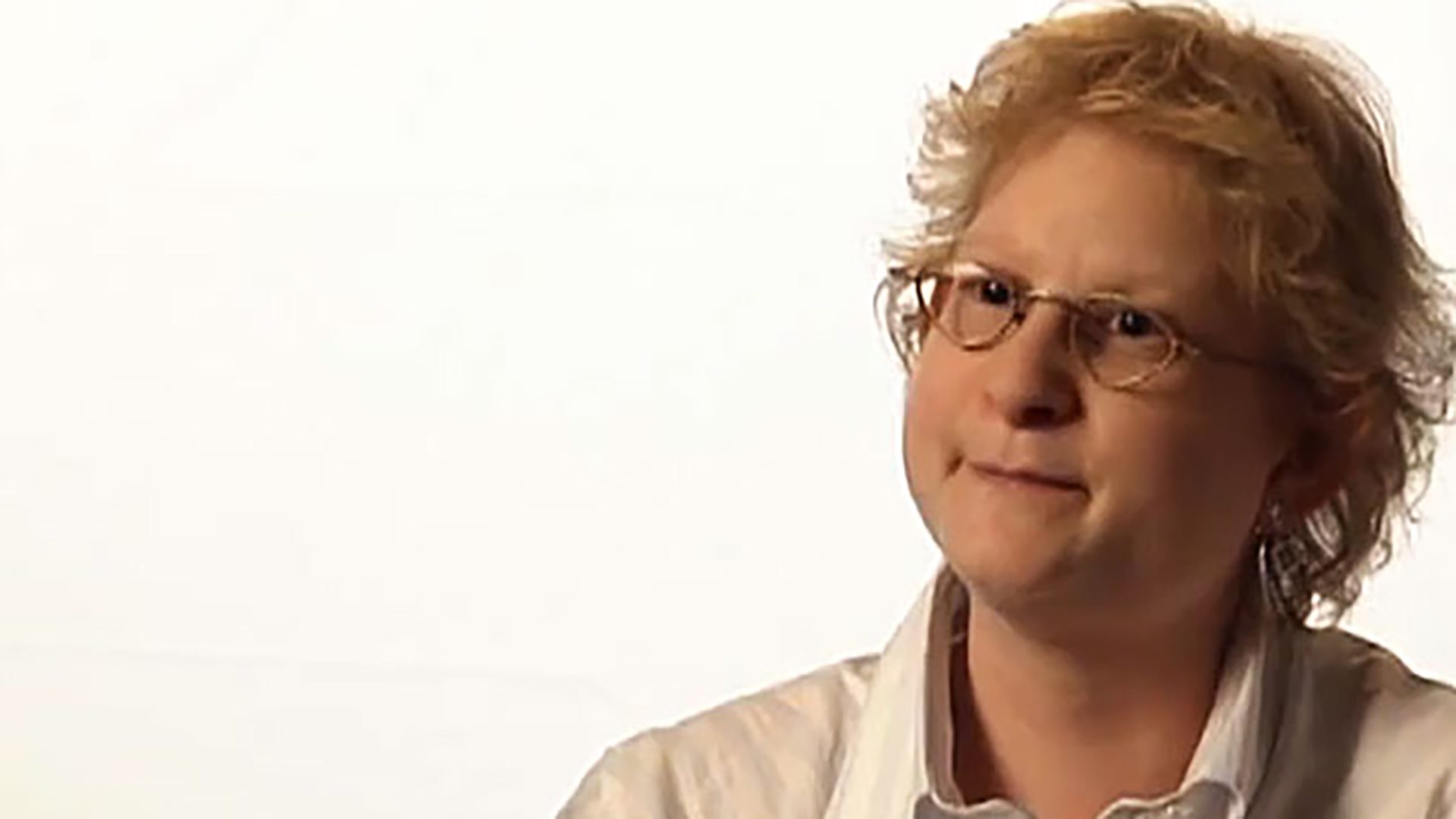 An adult woman with curly blonde hair and glasses wearing a white collared shirt is interviewed against a white background.