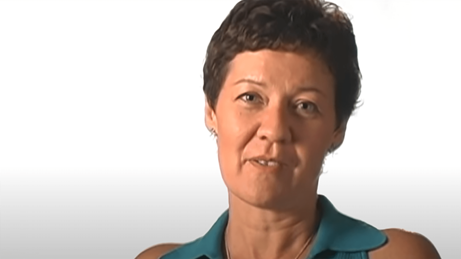 A middle aged woman with short hair and a teal shirt is interviewed against a white background