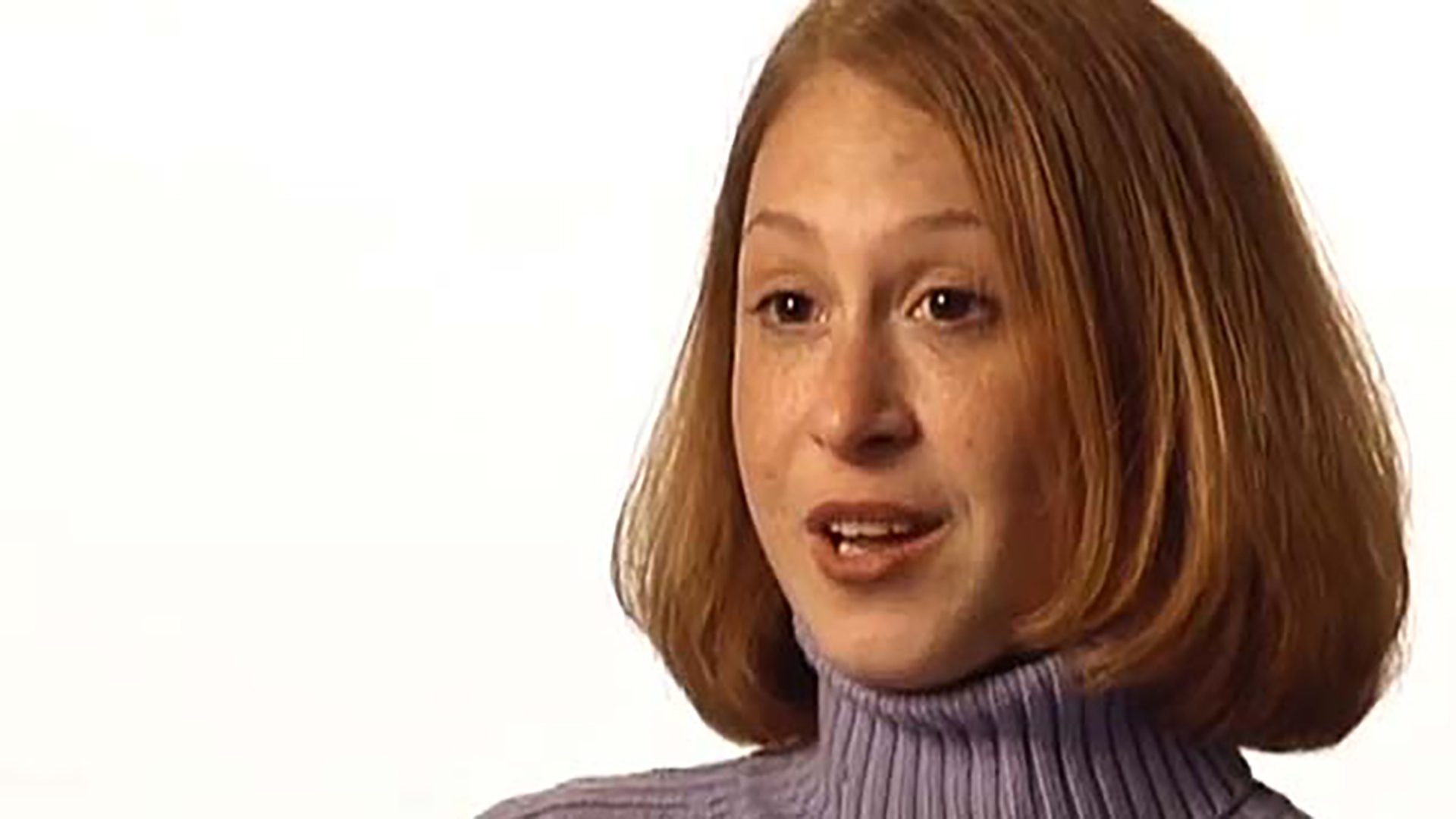 A young adult woman wearing a light purple turtleneck is interviewed against a white background.