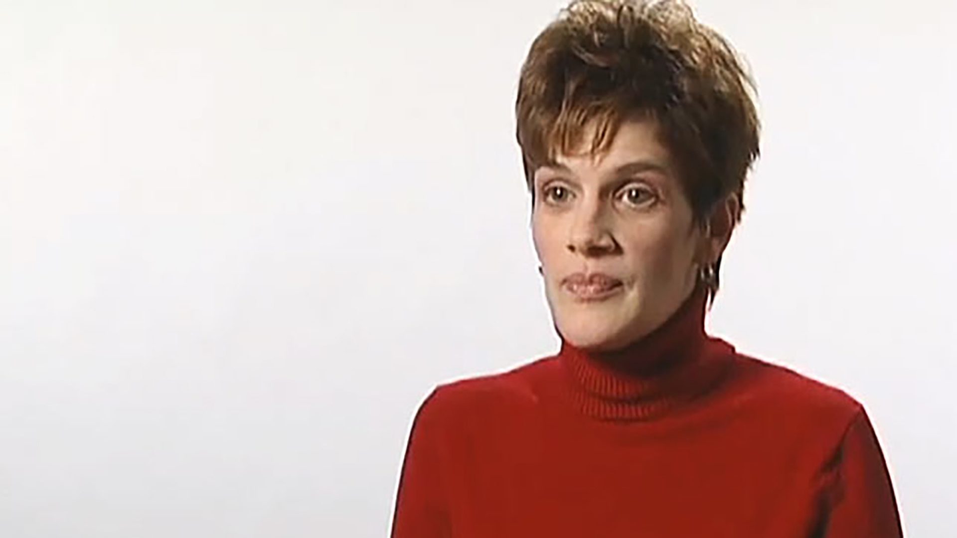 A woman with short hair wearing a red turtleneck is interviewed against a white background