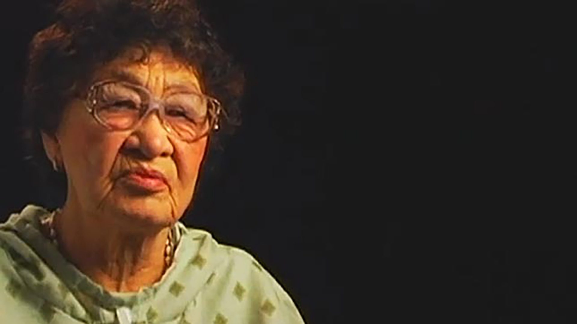 A senior woman with dark curly hair, glasses, and green patterned clothing is interviewed against a black background.