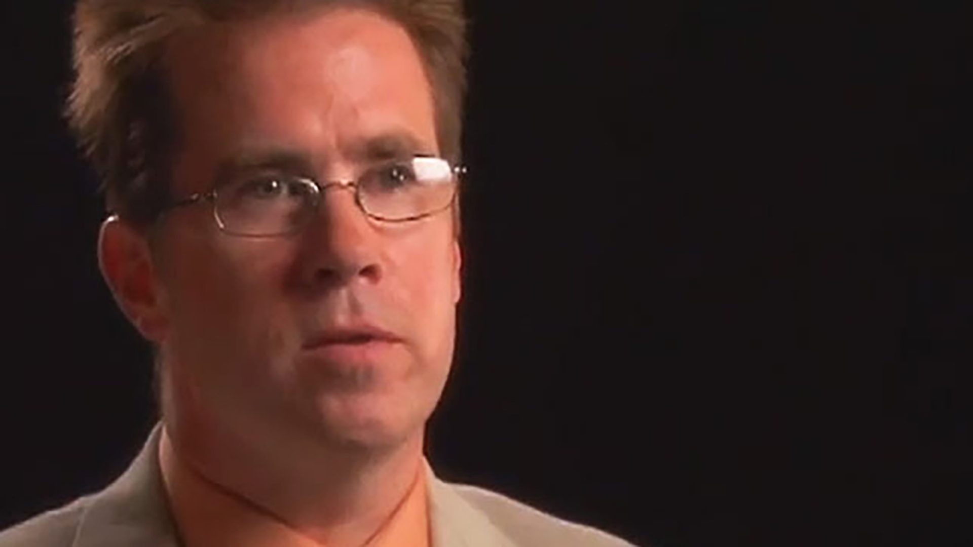 A man with glasses is interviewed against a black background