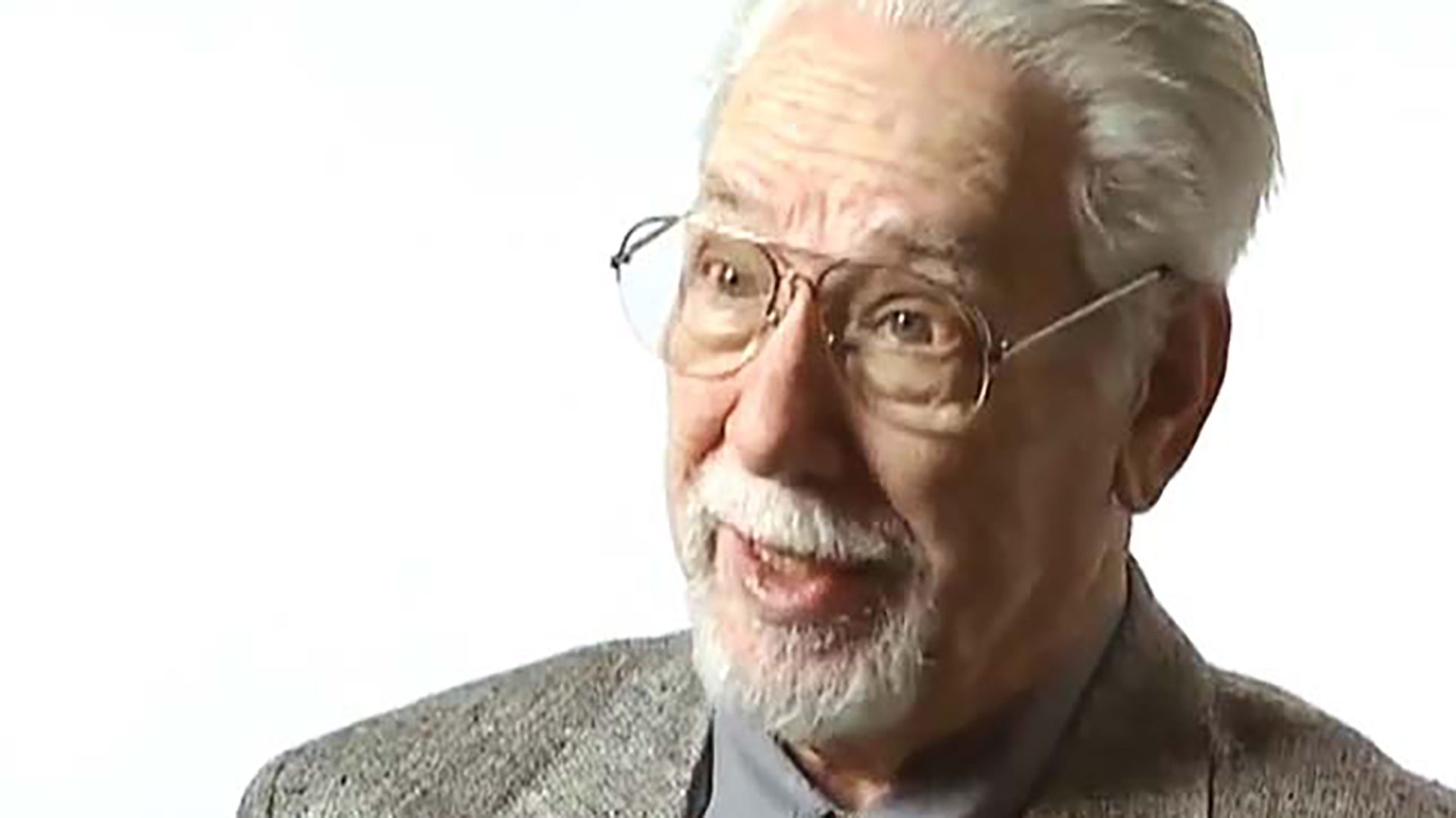 An expressive senior man with glasses and facial hair is interviewed against a white background.