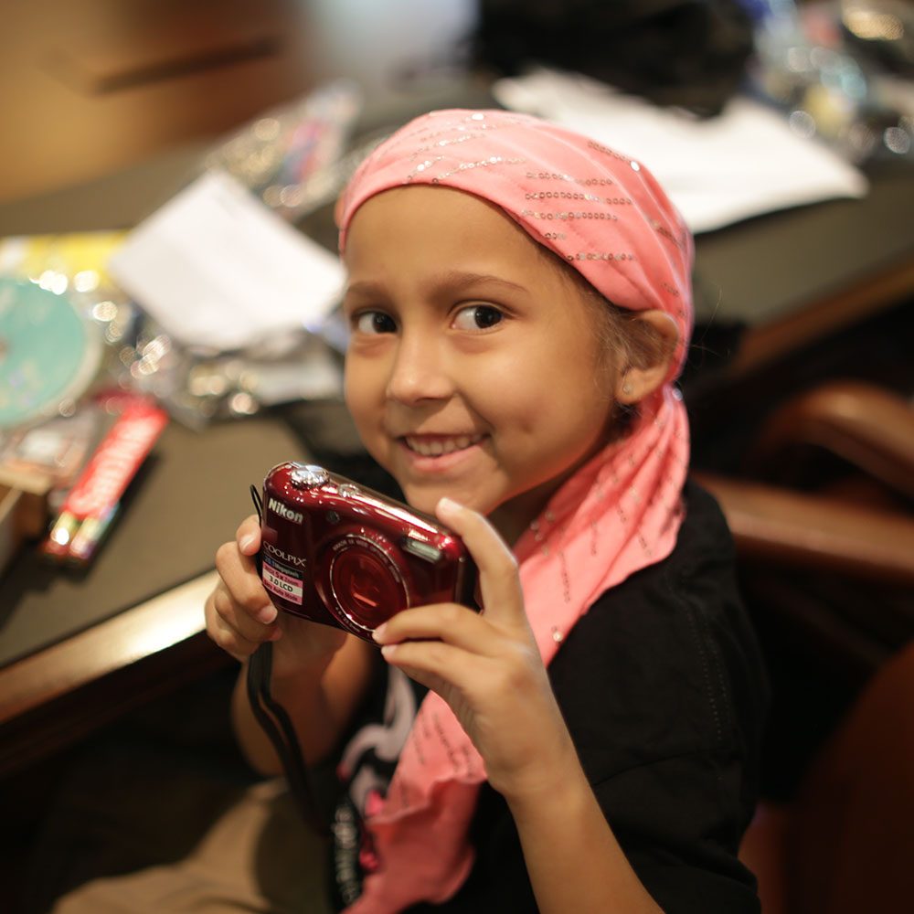 Pediatric cancer patient with pink head wrap holding camera