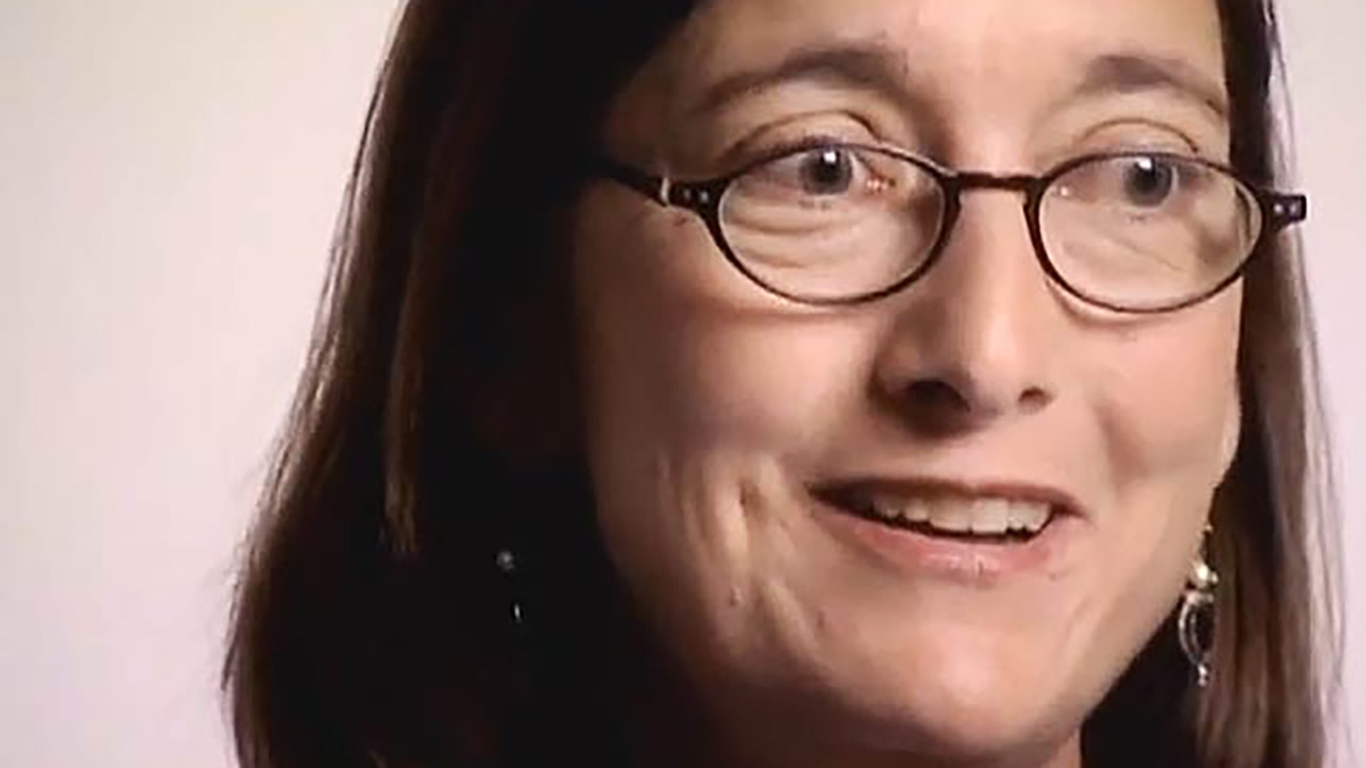 A close-up of an adult woman with dark hair and glasses being interviewed against a light background.