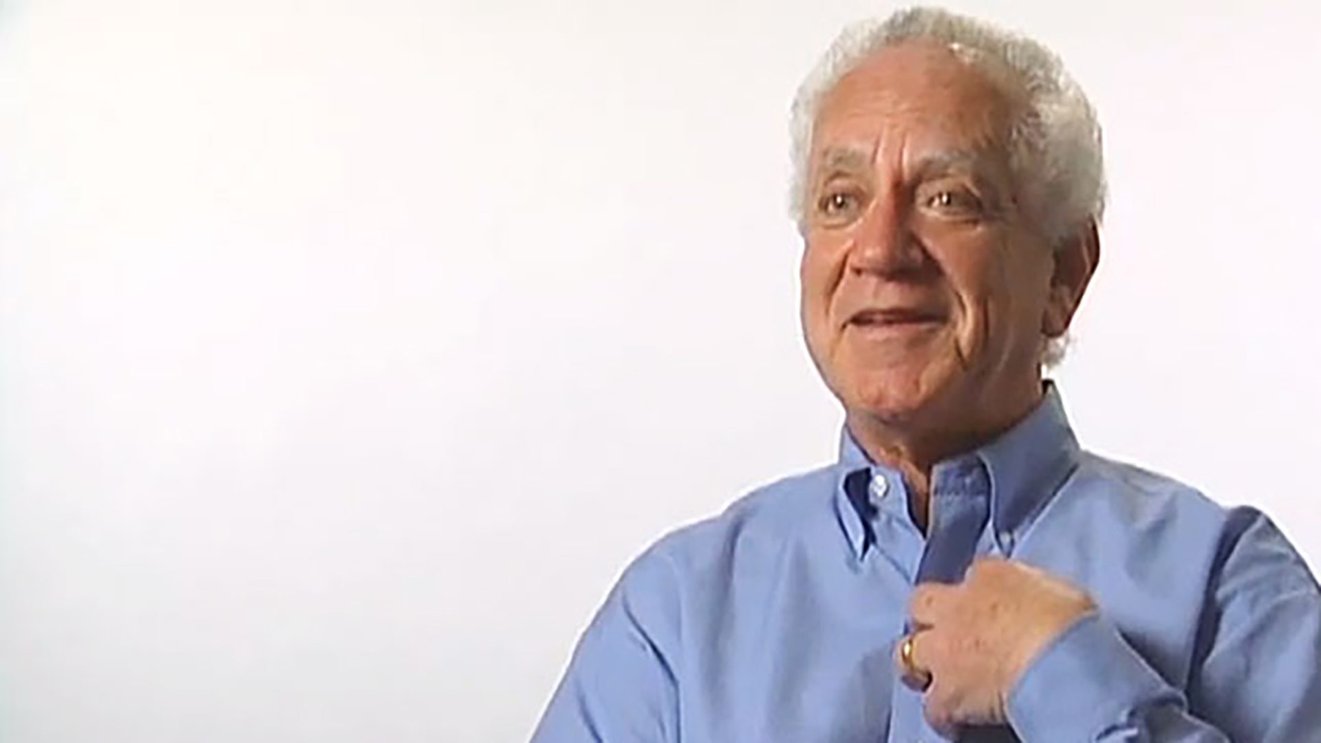 A senior man wearing a blue shirt is interviewed against a white background